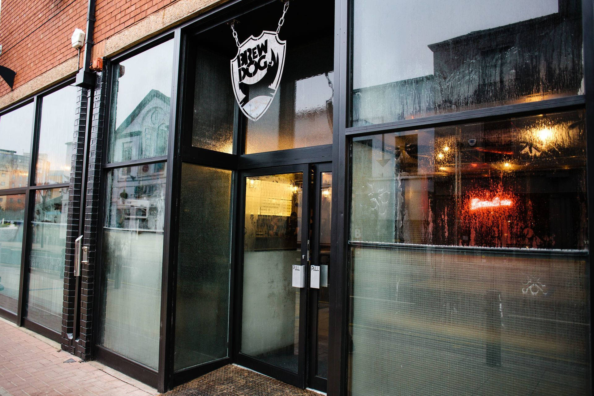 BrewDog 谢菲尔德分店外觀 壁紙 — 雨天的BrewDog酒吧入口景觀，濕漉漉的玻璃窗反射城市燈光，內部有發光的霓虹燈牌。