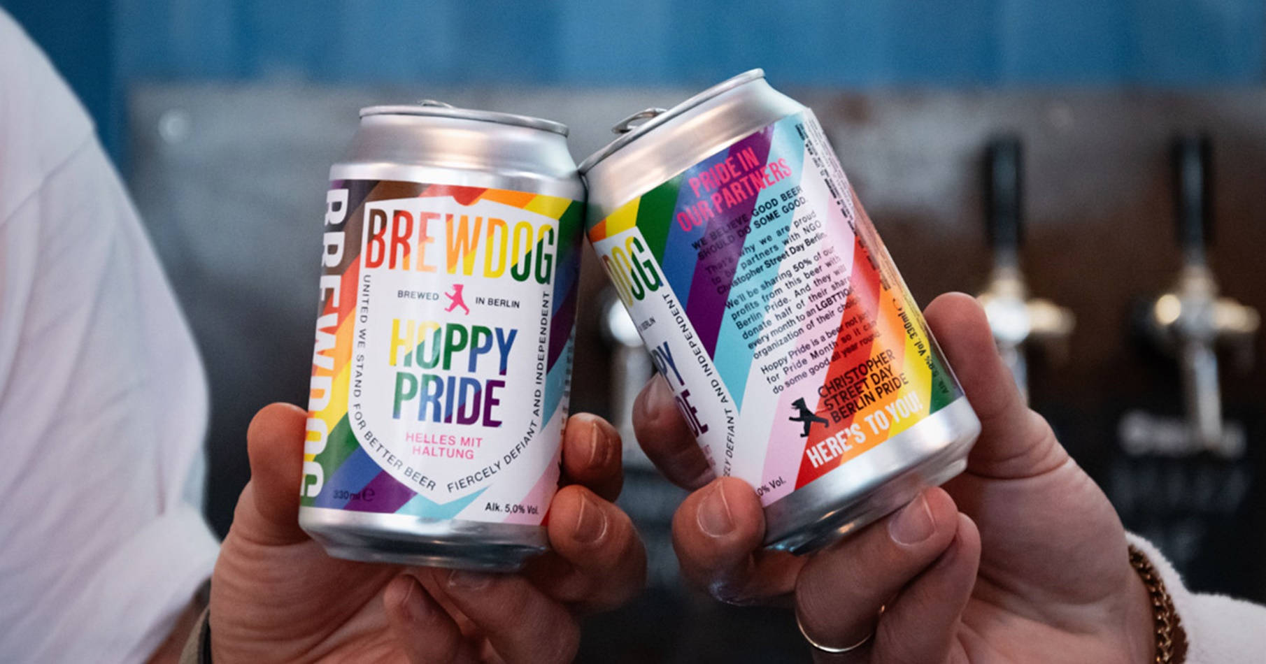 BrewDog Hoppy Pride 啤酒罐碰杯 壁紙 — 兩隻手在酒吧裡用彩虹主題的BrewDog啤酒罐碰杯，慶祝Pride。