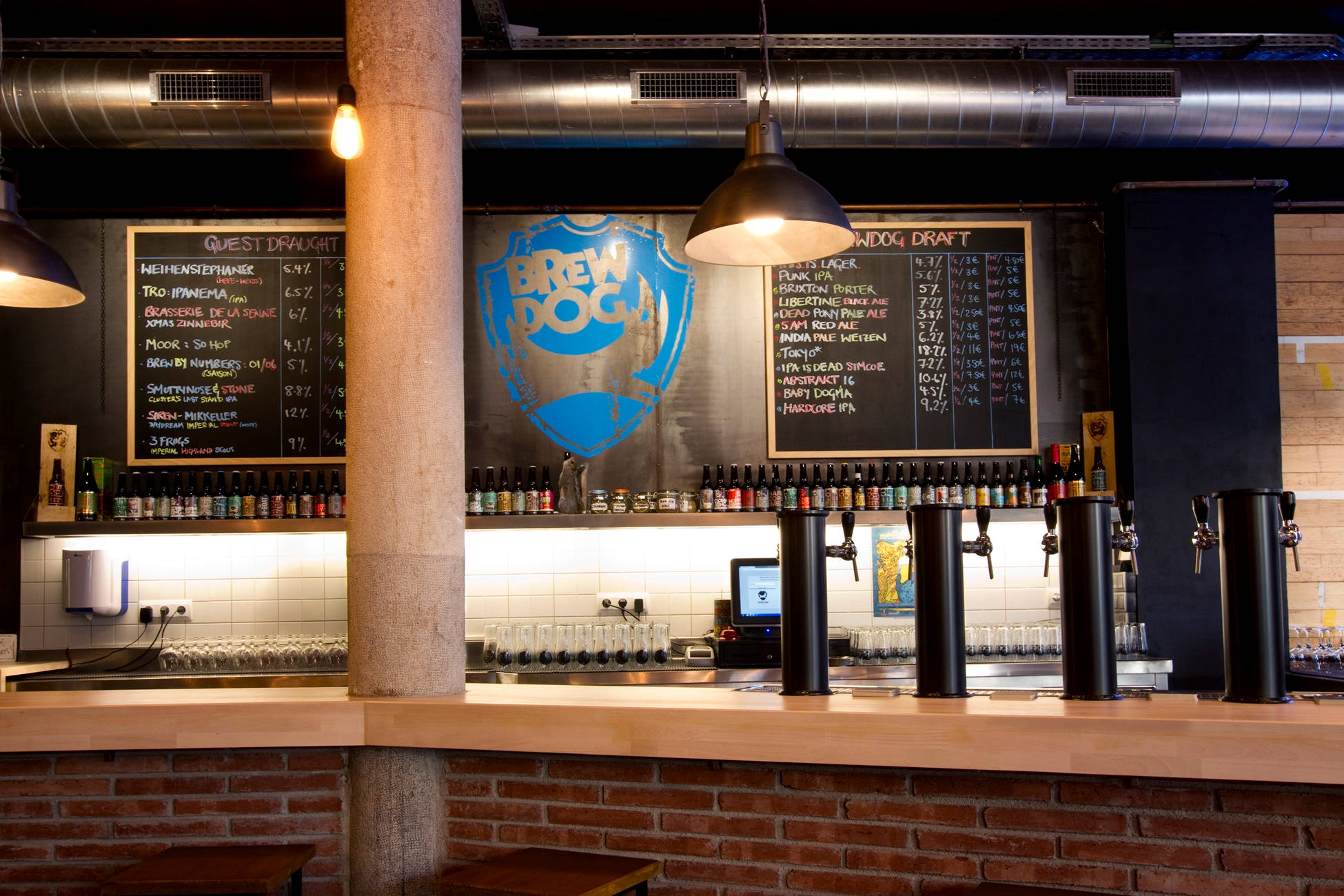 BrewDog 巴塞羅那分店內部 壁紙 — 燈光溫暖的BrewDog酒吧，有裸露磚牆、黑板菜單和掛在吊燈下的啤酒龍頭。