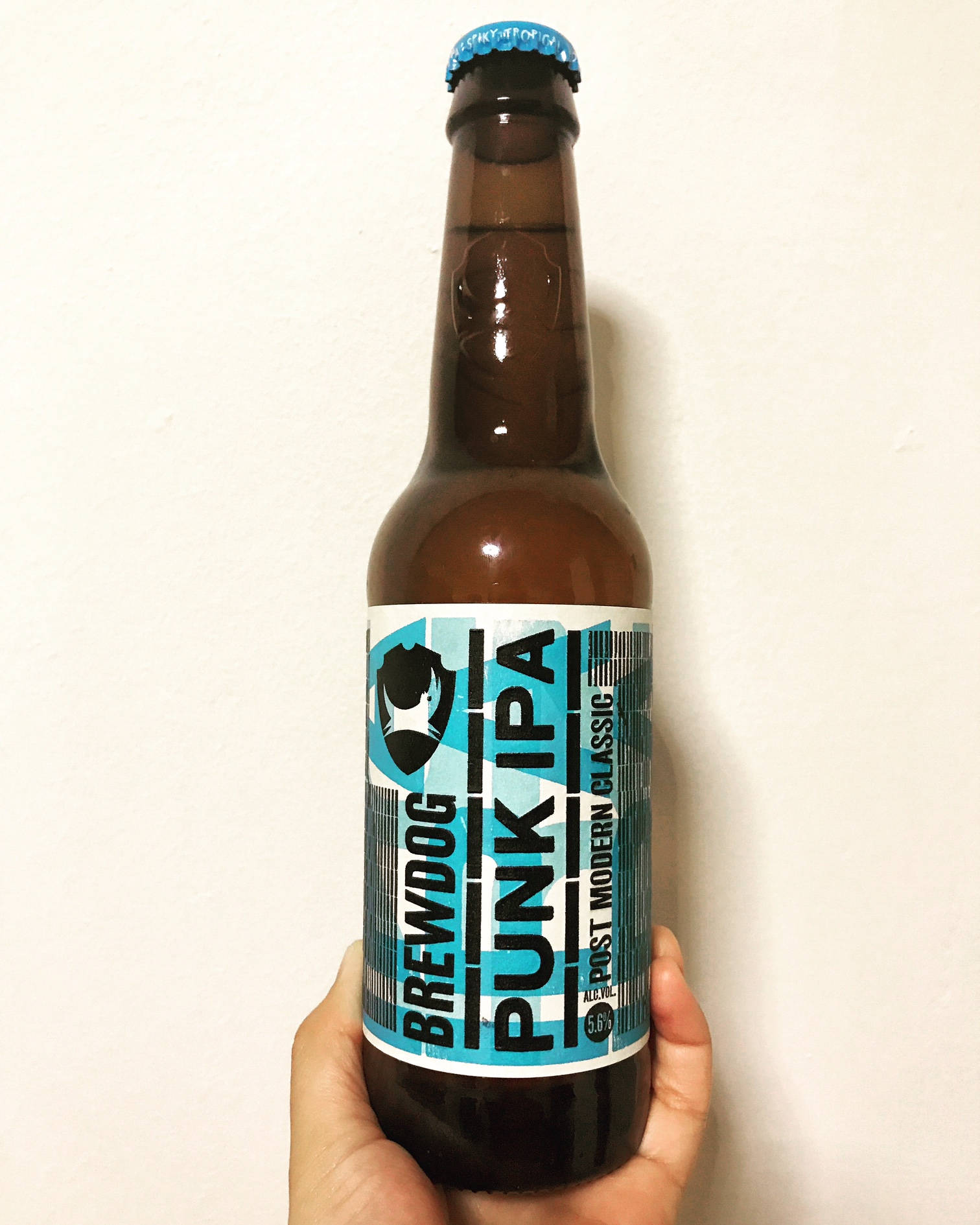 BrewDog Punk IPA 啤酒瓶 壁紙 — 一隻手拿著一個棕色 BrewDog Punk IPA 啤酒瓶，對著白色素牆，瓶蓋為藍色，標籤大膽。