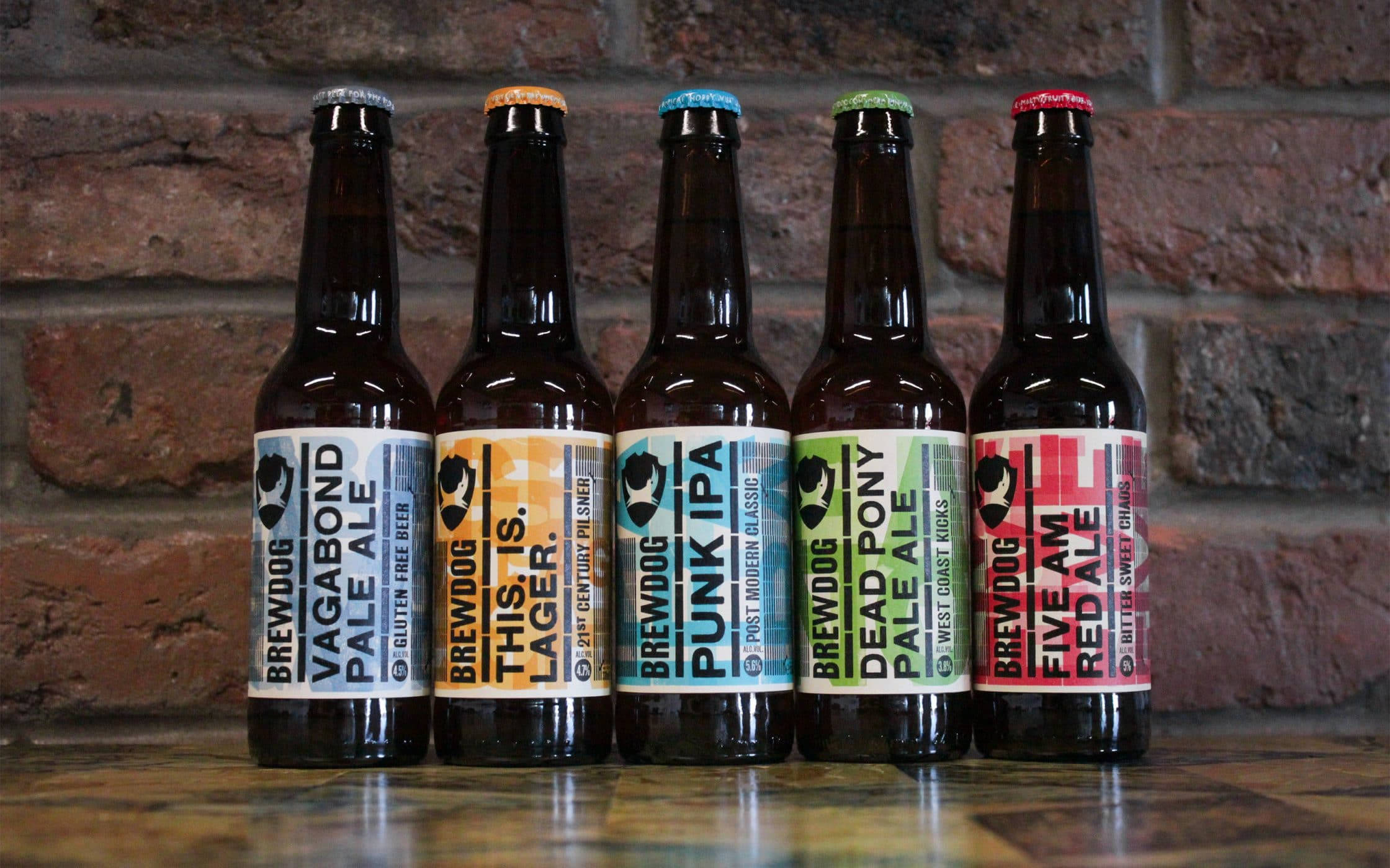 BrewDog 精釀啤酒瓶排列於磚牆前 壁紙 — 五款色彩繽紛的BrewDog啤酒瓶排列在木質表面上，背景是質樸的磚牆。