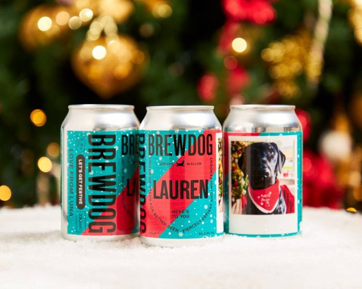 BrewDog Trio Delight - 三款不同啤酒罐 壁紙 — 三個個人化的BrewDog啤酒罐，帶有節日設計，放在雪地上，背景是模糊的聖誕樹燈光。