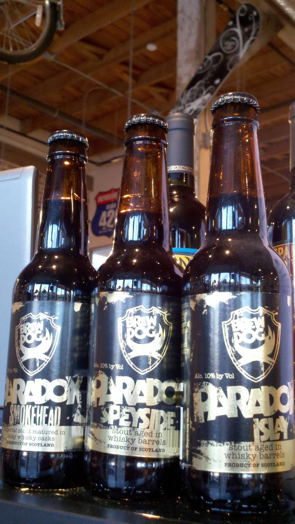 BrewDog 三瓶 Paradox Islay 壁紙 — BrewDog Paradox 黑啤酒玻璃瓶的特寫，排列在木樑下的架子上，上方有一個自行車輪。