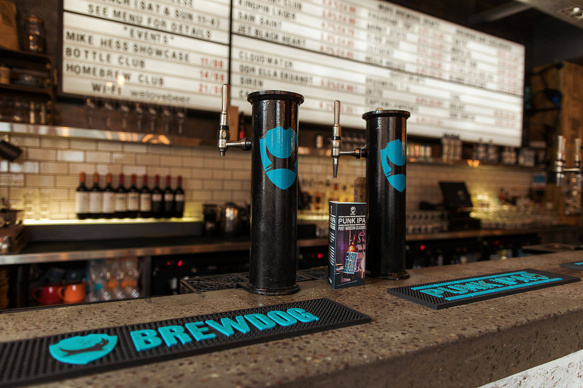 BrewDog 坎登酒吧熱鬧夜景 壁紙 — 兩個帶有藍色標誌的黑色啤酒龍頭，放在一個有斑點的酒吧枱面上，配有品牌杯墊和背後發光的餐牌板。