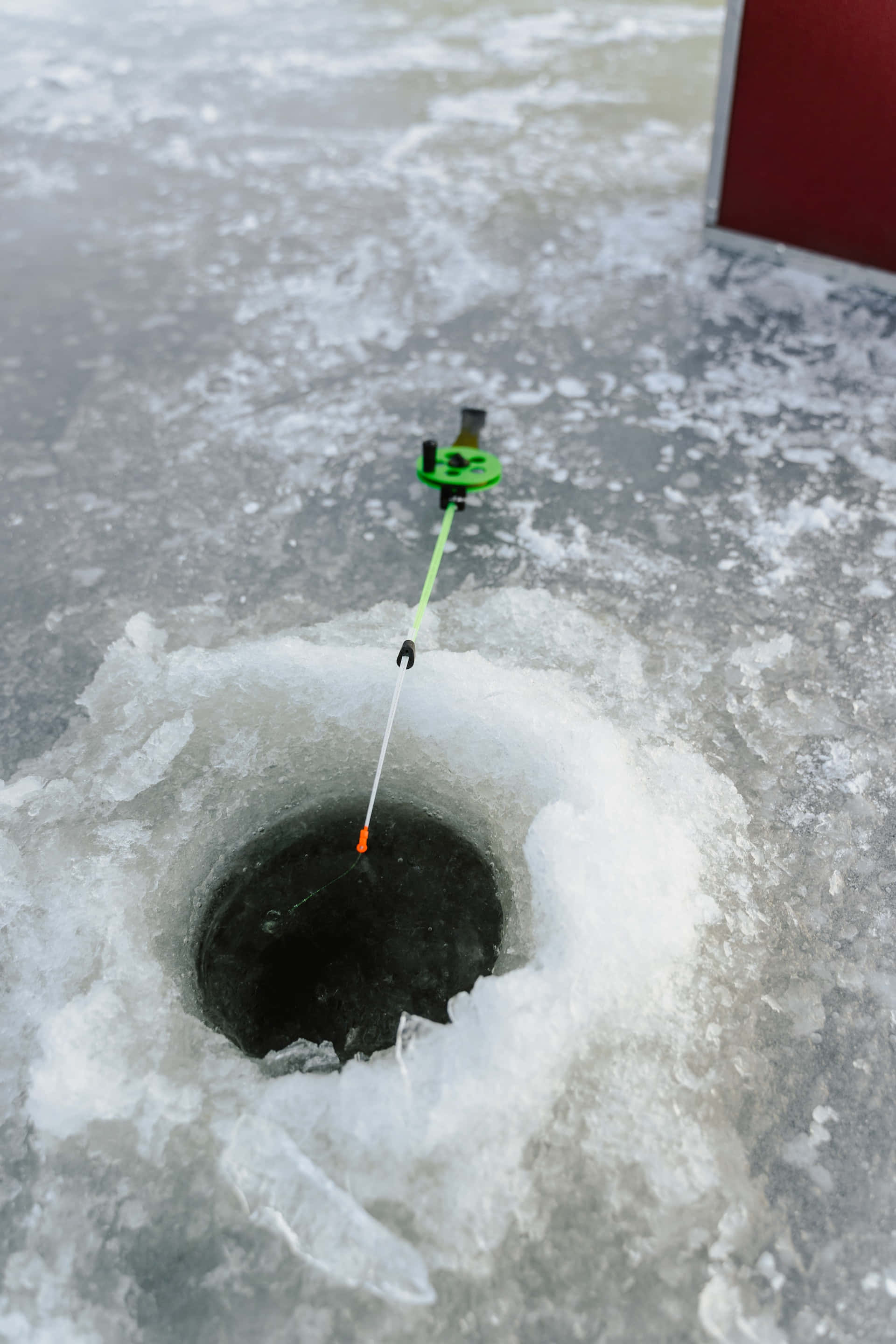 Ice Fishing 桌布 壁紙 — 一個鮮綠色的冰釣提示器在陰天之下，立於一個紋理冰凍湖面的暗洞之上。