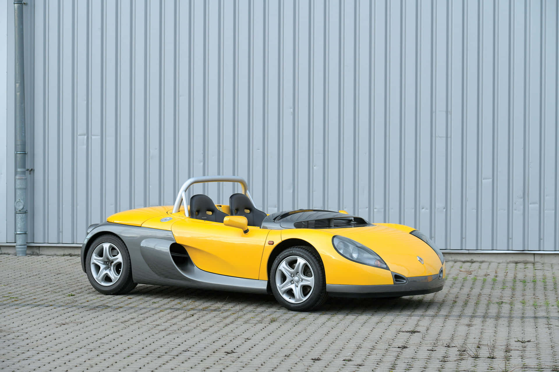 Renault Sport Spider 桌布 壁紙 — 一輛亮黃色和銀色的雙座跑車停泊在鋪砌路面上，背景是波紋金屬牆。