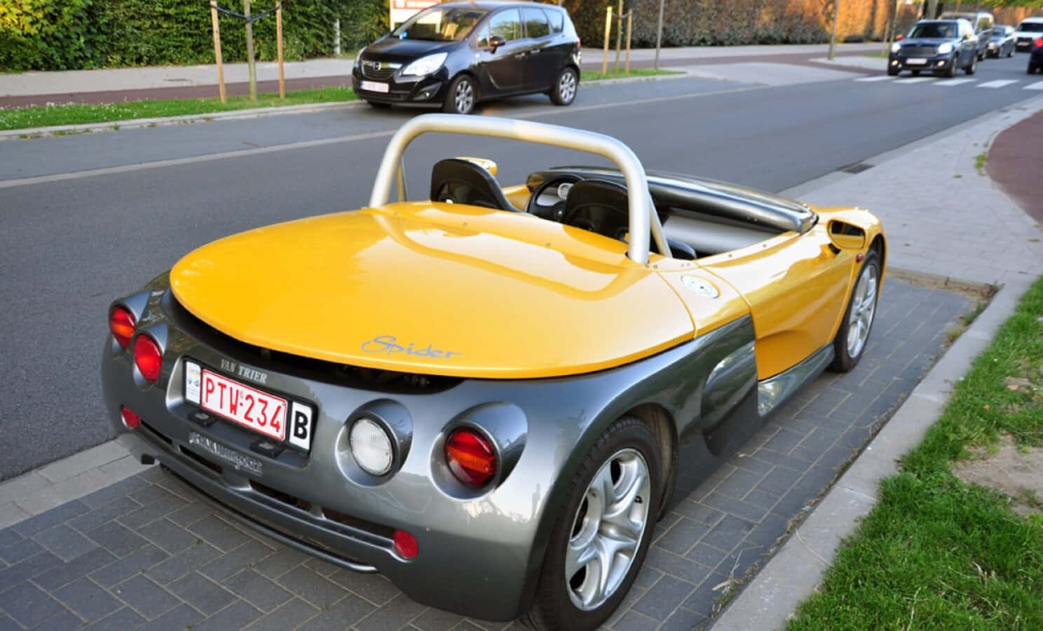 Renault Sport Spider 桌布 壁紙 — 一輛亮黃色和銀色的TVR Spider開篷車停泊在磚砌行人路上，旁邊是一條有其他車輛和綠化的街道。