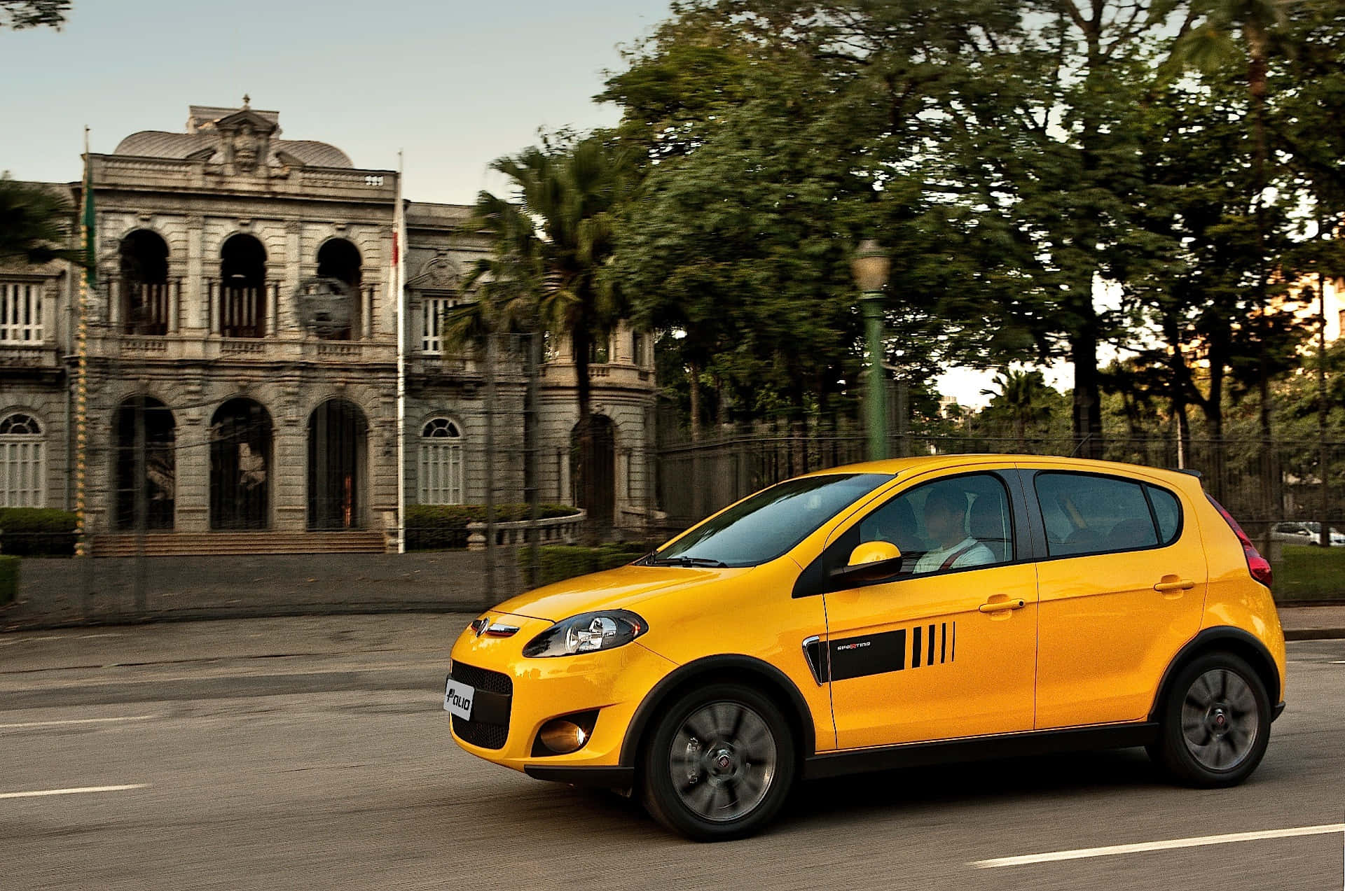 Fiat Palio 桌布 壁紙 — 一輛亮黃色的快意Uno駛過一座帶有拱形窗戶和茂密綠樹的宏偉石建築。