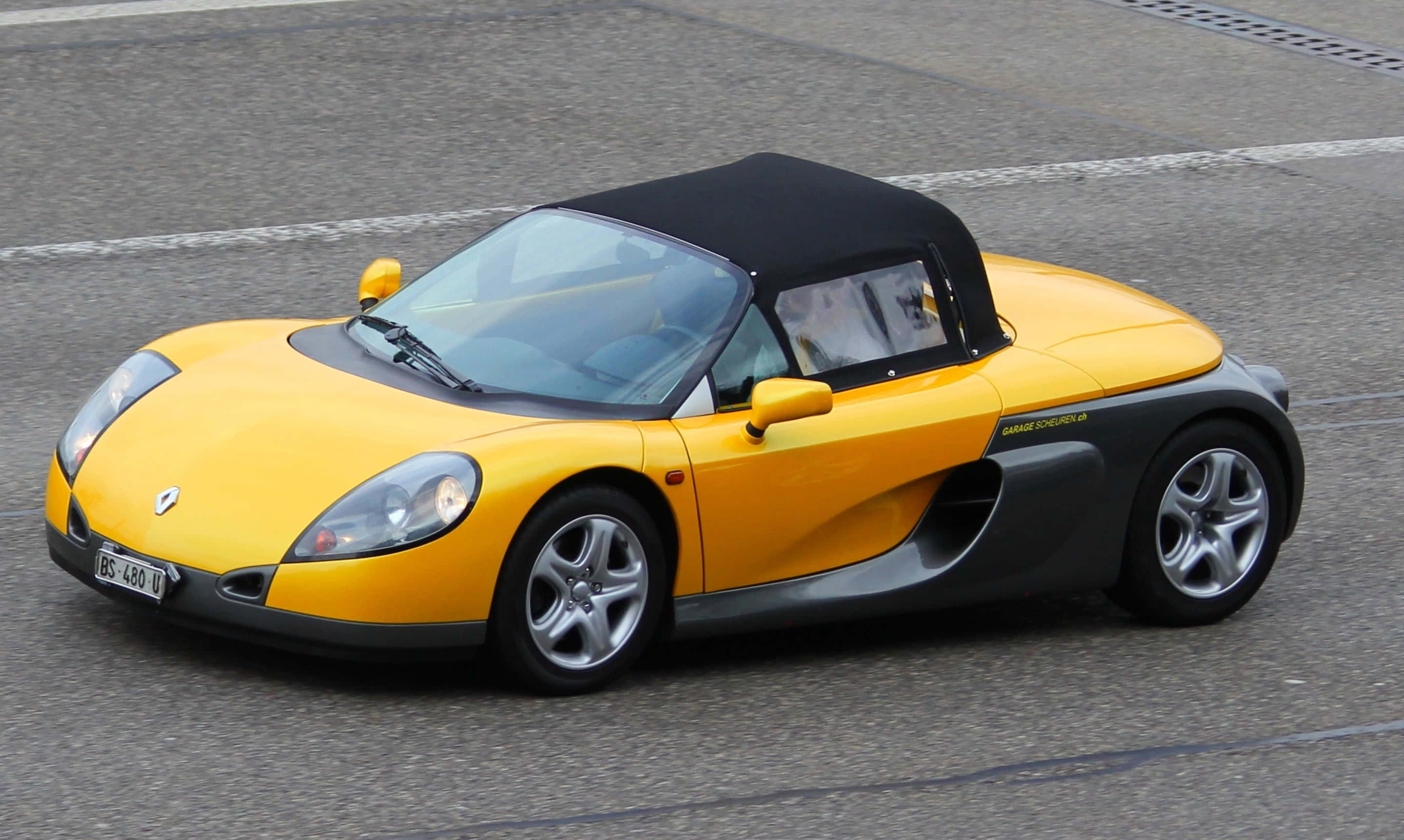 Renault Sport Spider 桌布 壁紙 — 一輛亮黃色雷諾Spider開篷車，配備黑色軟頂，停泊在灰色瀝青路面上。