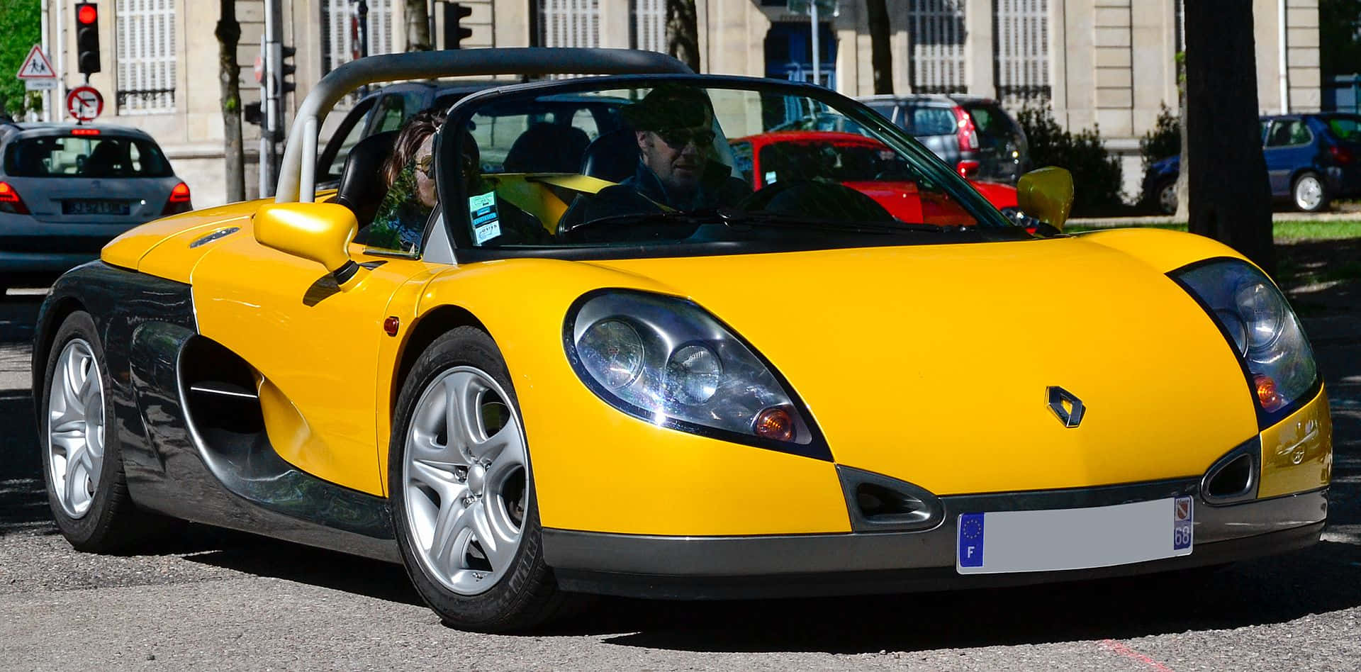 Renault Sport Spider 桌布 壁紙 — 一輛亮黃色的雷諾Spider跑車停泊在陽光明媚的城市街道上，車內有人。