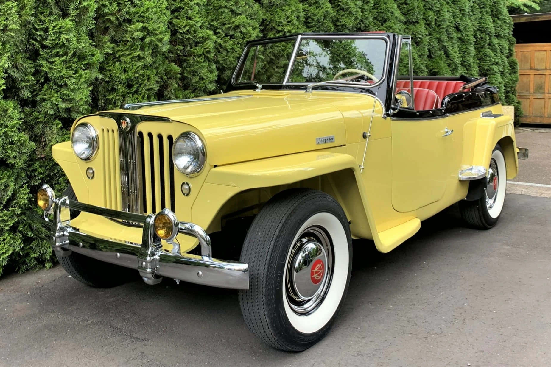 Jeepster 桌布 壁紙 — 一輛亮黃色復古敞篷車，紅色內飾，停在鋪砌車道旁茂密的綠色樹籬邊。