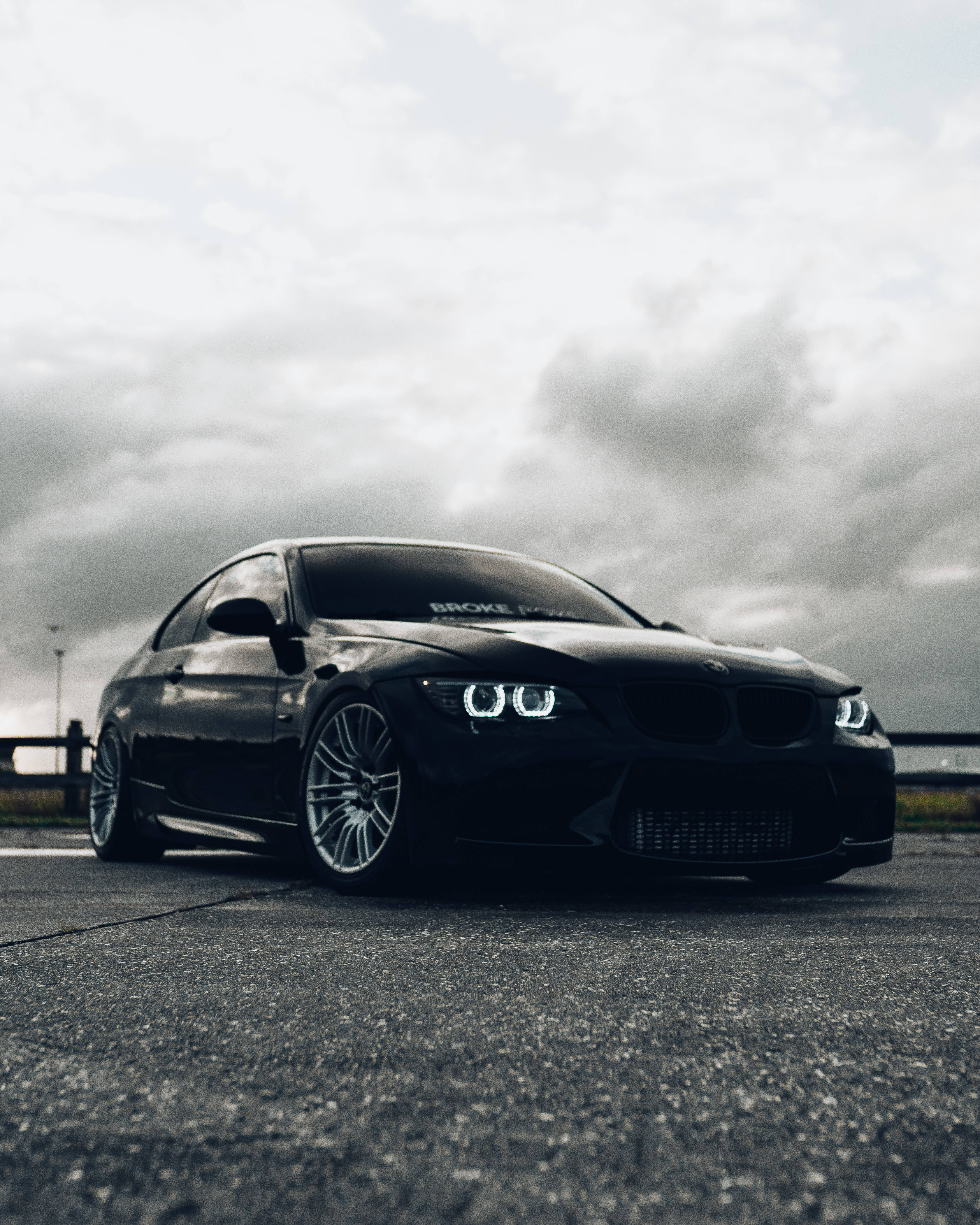 《Broke Boys》黑色寶馬 M3 壁紙 — 一輛時尚的黑色BMW跑車，配備發光的車頭燈，停在多雲天空下的瀝青路面上。