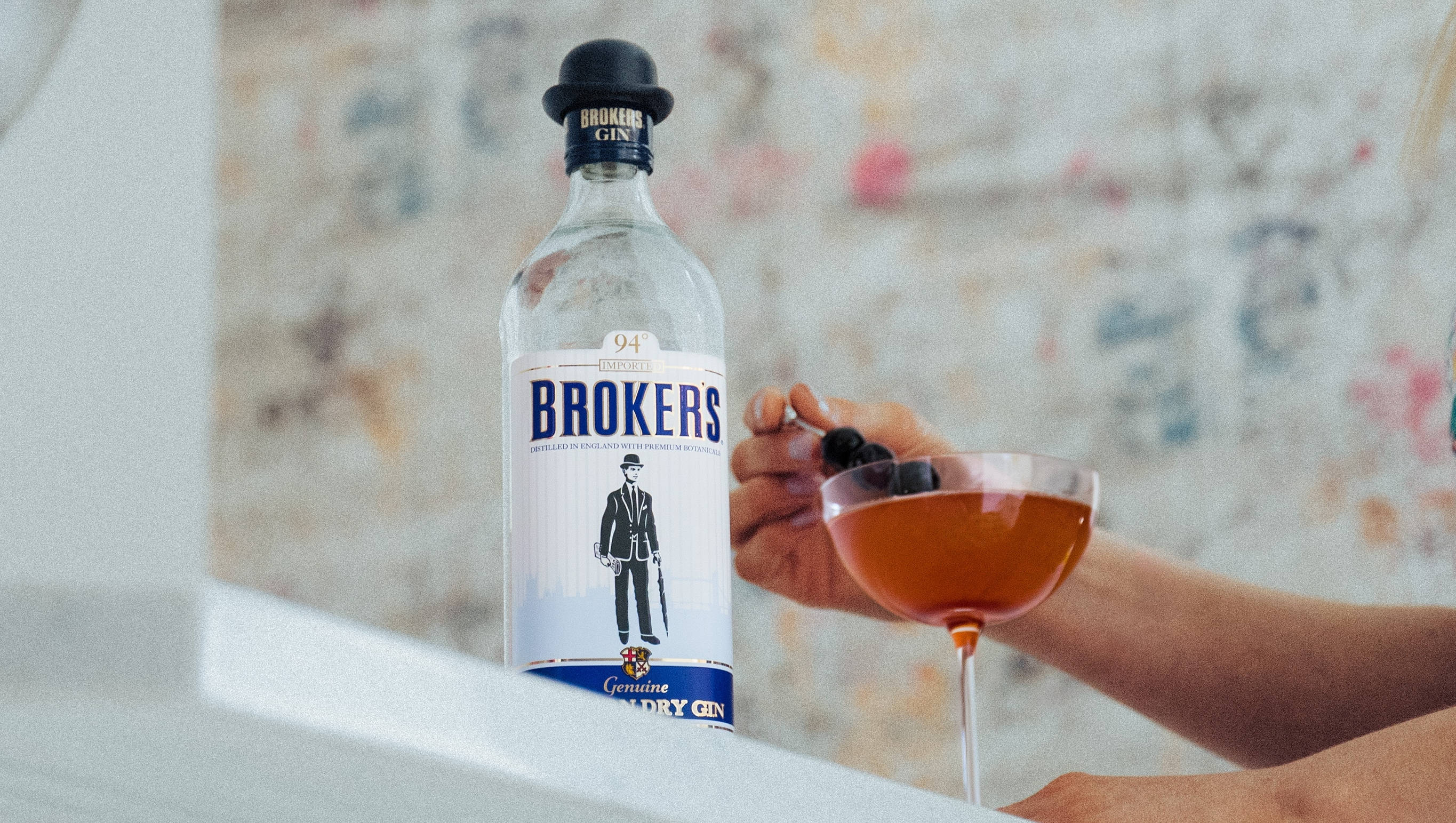 高雅 Broker's 金酒曼哈頓雞尾酒 壁紙 — 一隻手在白色表面上拿著一杯裝有琥珀色液體的雞尾酒杯，旁邊放著一瓶Brokers氈酒，背景是花卉牆紙。