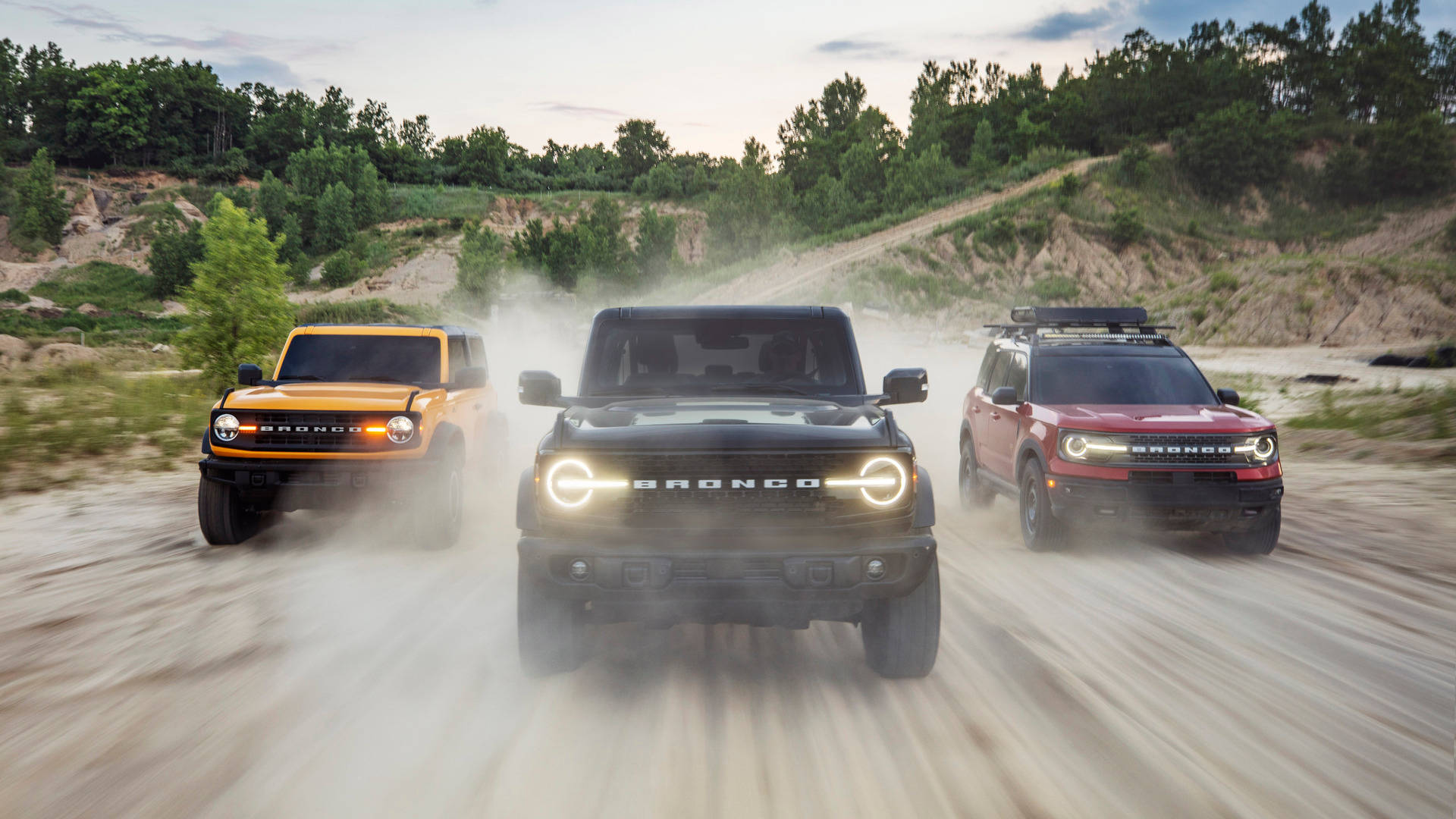 三輛不同顏色的福特 Bronco 壁紙 — 三輛橙色、黑色和紅色的福特 Bronco 在塵土飛揚的小路上咆哮，揚起滾滾塵土，背景是樹木繁茂的山坡。