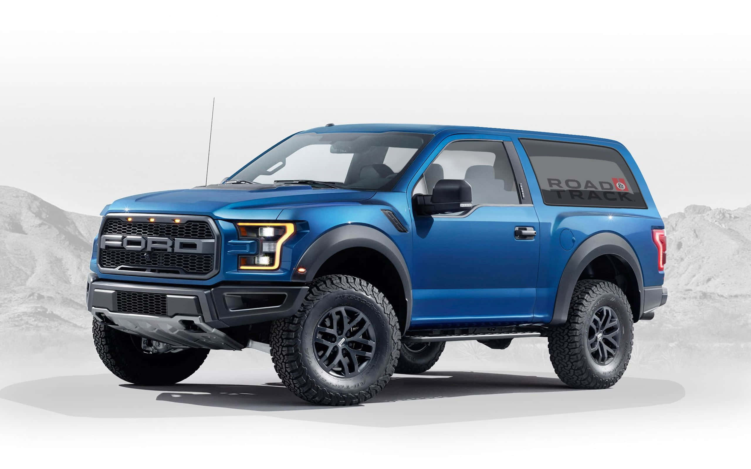 堅韌時尚的福特 Bronco 壁紙 — 一輛充滿活力的藍色福特 F-150 Raptor SUV，帶有黑色飾邊和「ROAD TRACK」貼花，在荒涼的山景背景下蓄勢待發。
