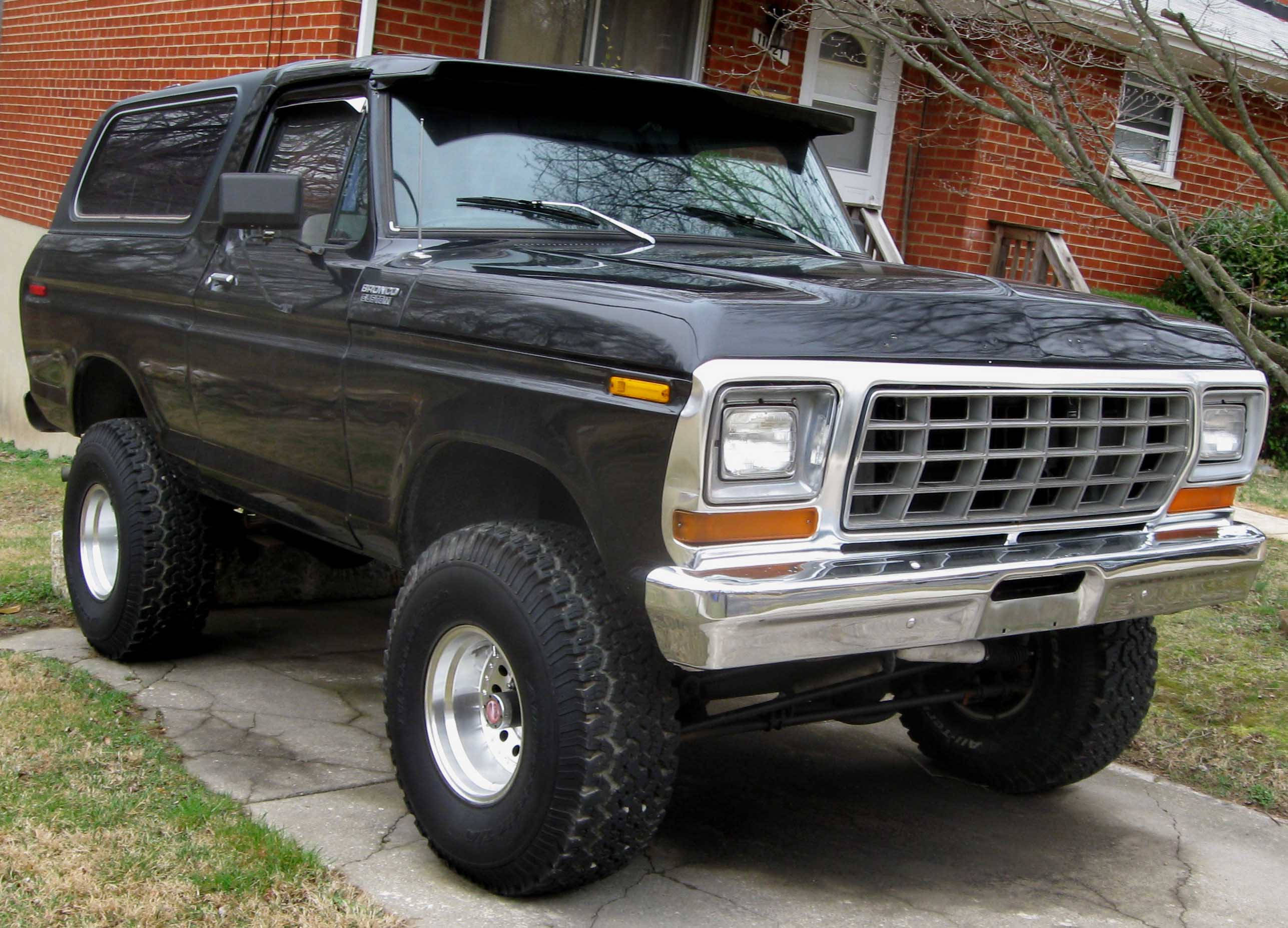 福特Bronco鍍鉻前格柵 壁紙 — 一輛黑色升高版福特Bronco SUV，配備鍍鉻格柵和大輪胎，停在紅磚屋前裂開的混凝土車道上。