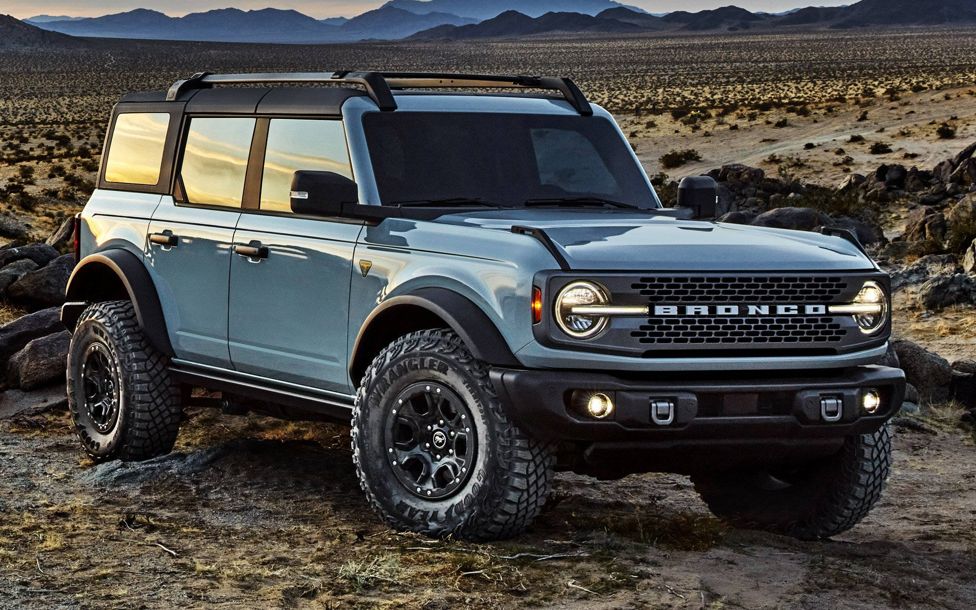 福特 Bronco 停在荒蕪道路 壁紙 — 一輛淺藍色福特Bronco SUV，帶有黑色飾邊和堅固輪胎，在金色日落天空和遠山下的岩石沙漠地形上蓄勢待發。