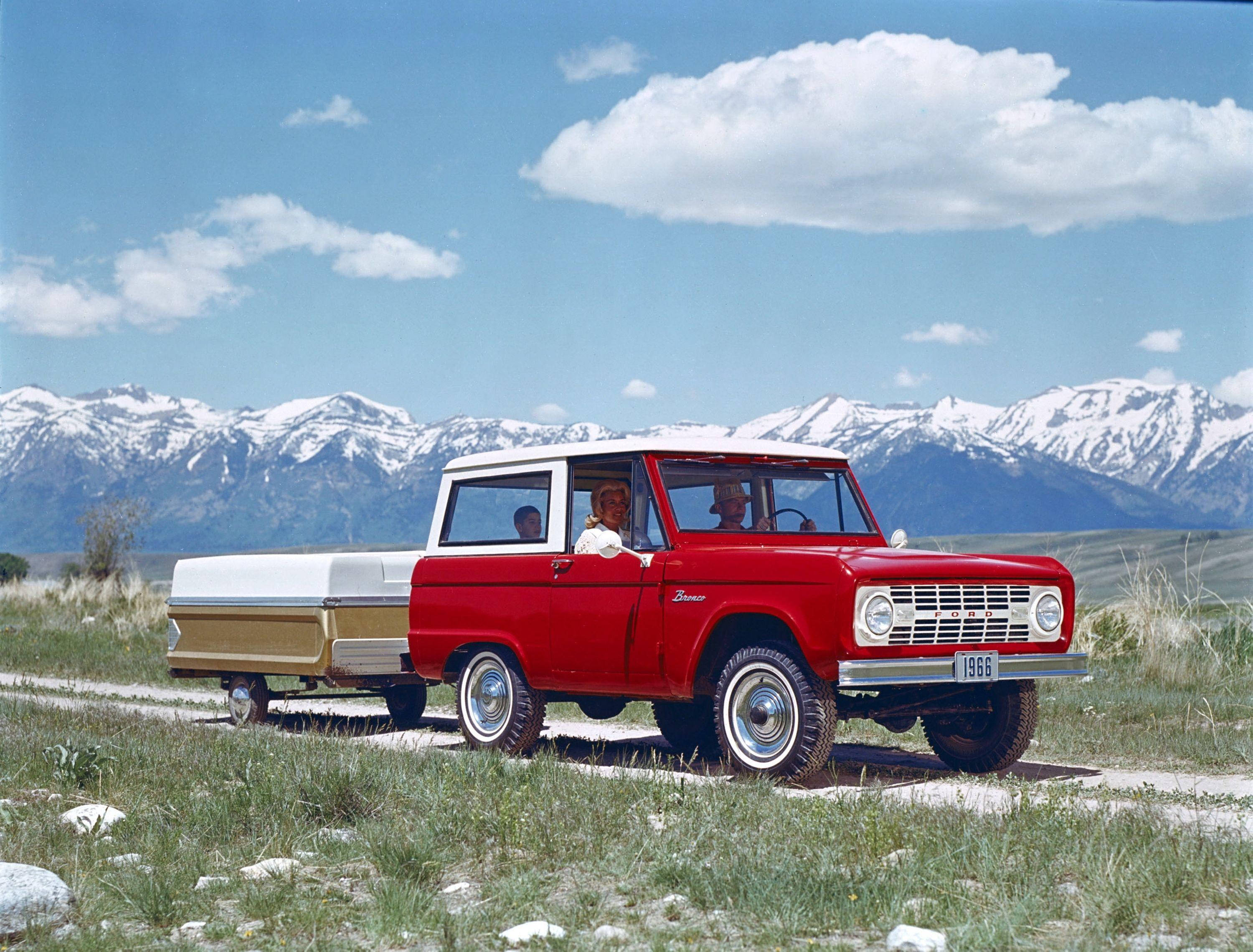 紅白雙色福特 Bronco 與拖車 壁紙 — 一輛復古紅白相間的1966年福特Bronco在藍天白雲下，駛過一片草地，遠處是白雪覆蓋的山峰。