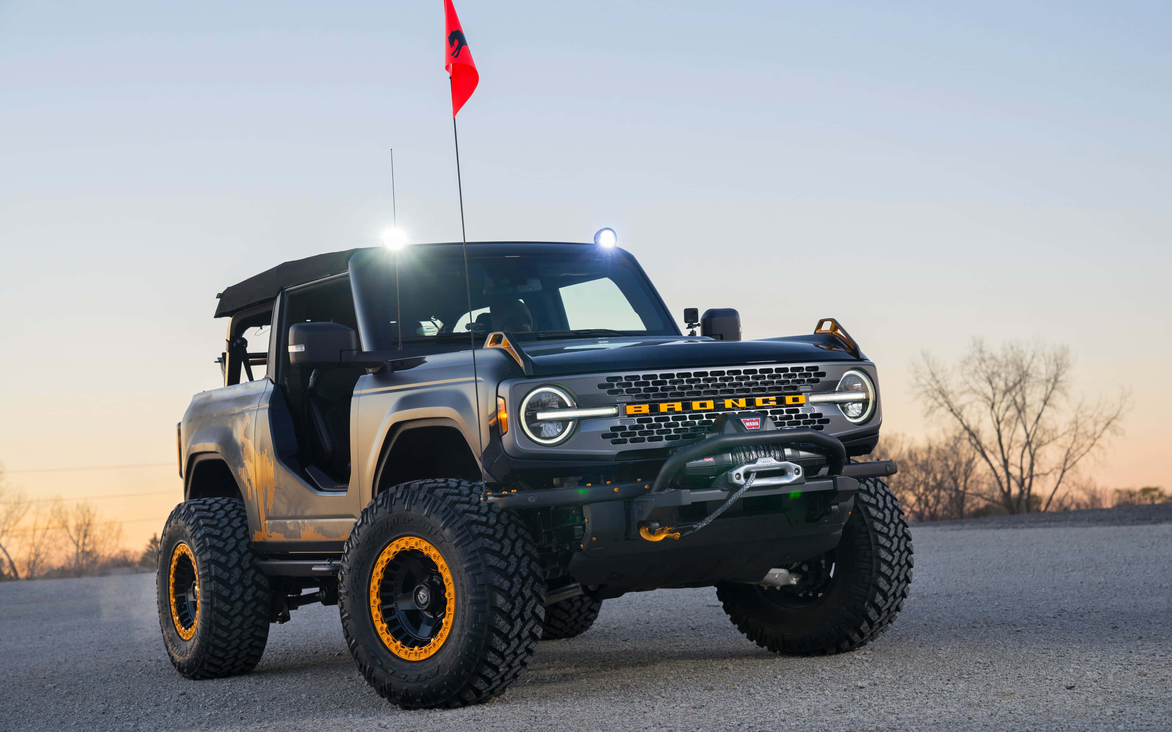 福特Bronco — 准備迎接任何冒險 壁紙 — 一架堅固的黑色福特Bronco，帶有橙色車輪點綴，在日落時分停在鋪砌的道路上，背景是光禿禿的樹木。