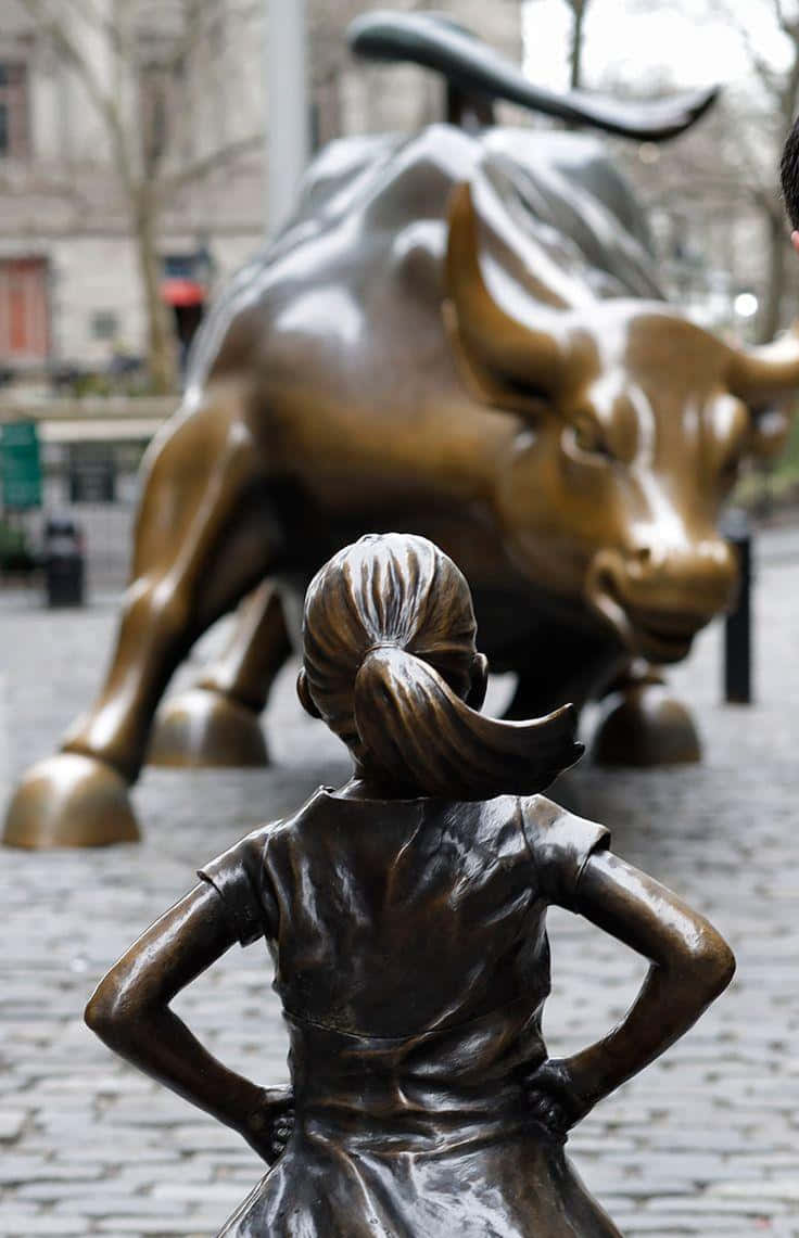 Bull Market 桌布 壁紙 — 一個雙手叉腰的女孩銅像，在城市廣場上挑釁地站在衝鋒牛雕像前。