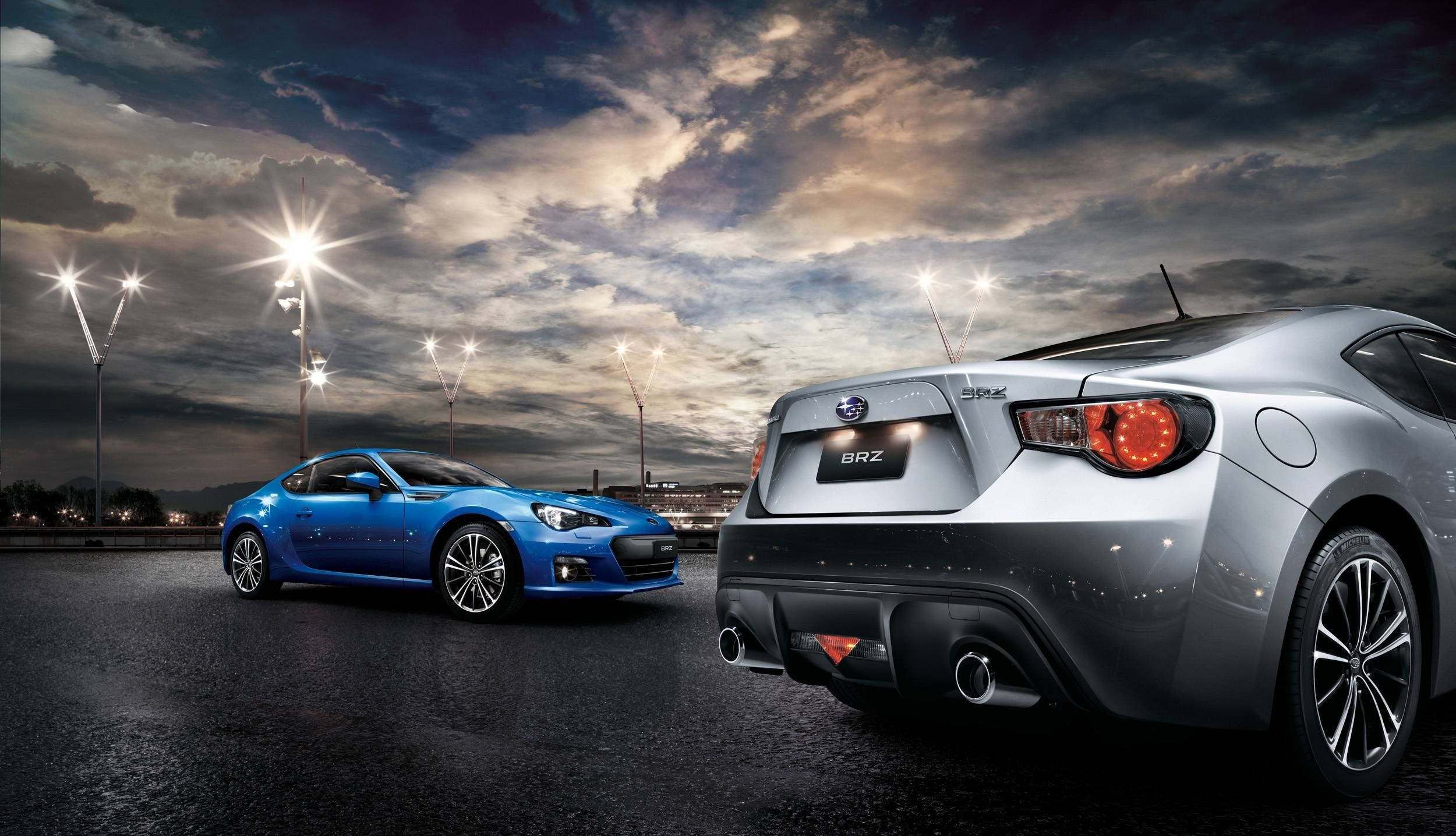 燈光璀璨的BRZ 壁紙 — 兩輛時尚的Subaru BRZ跑車，一藍一銀，停在潮濕的賽車場上，背景是戲劇性的暮色天空和閃爍的體育場燈光。