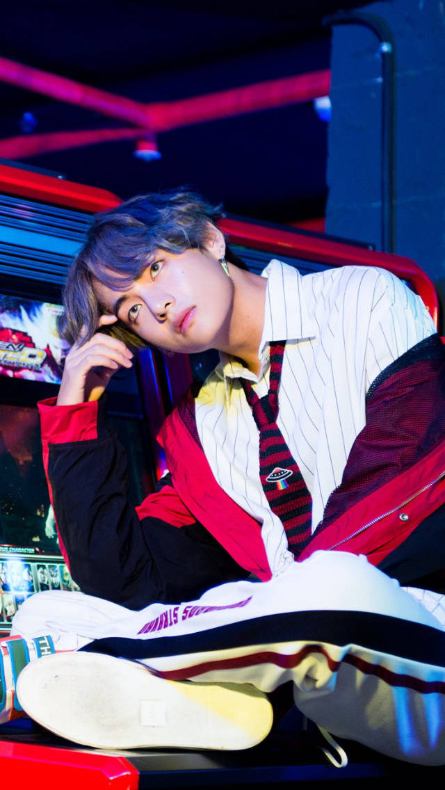 BTS 田柾國在遊戲機廳 壁紙 — 一位藍髮年輕男子在霓虹燈閃爍的遊戲廳中悠閒地休息，穿著紅黑相間的夾克，內搭條紋襯衫。