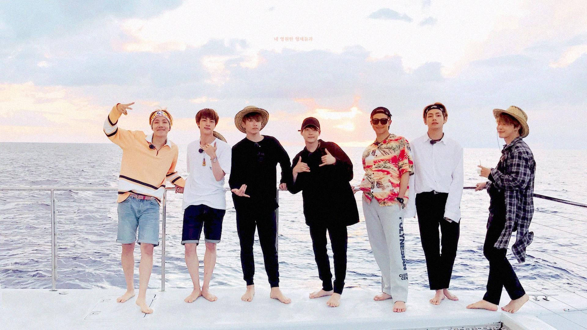 BTS 2020 船上休閒 壁紙 — 七位身穿休閒夏日服裝的男士赤腳在船甲板上擺姿勢，背景是柔和的日落天空和寧靜的海洋。