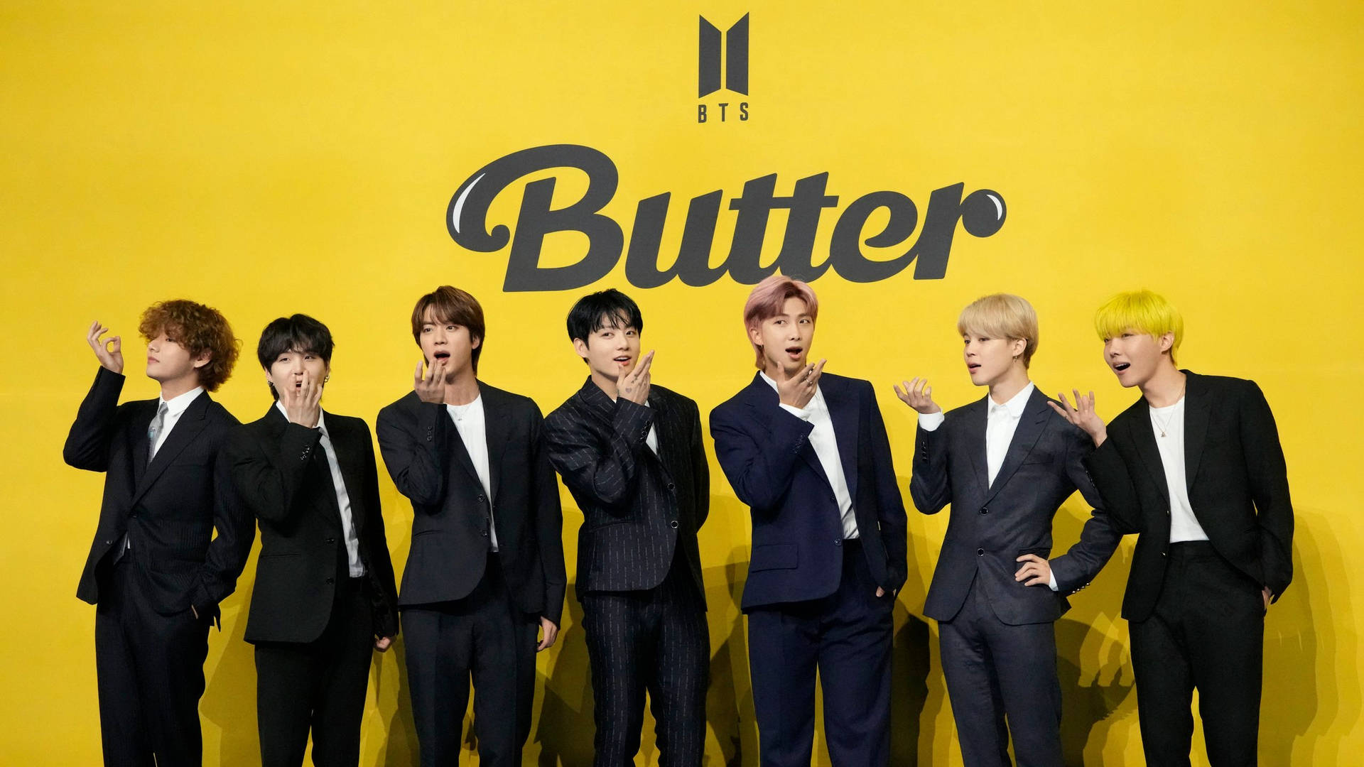 七名身穿黑色西裝的 K-pop 偶像在黃色 'Butter' BTS 背景前擺姿勢。