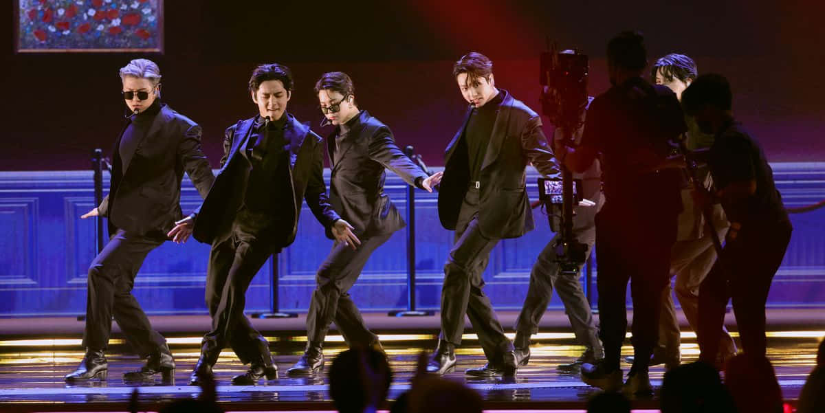 BTS 2022《Butter》表演現場照片 壁紙 — 五位身穿黑色西裝的男舞者在戲劇性的紅色和藍色舞台燈光下表演同步編舞。