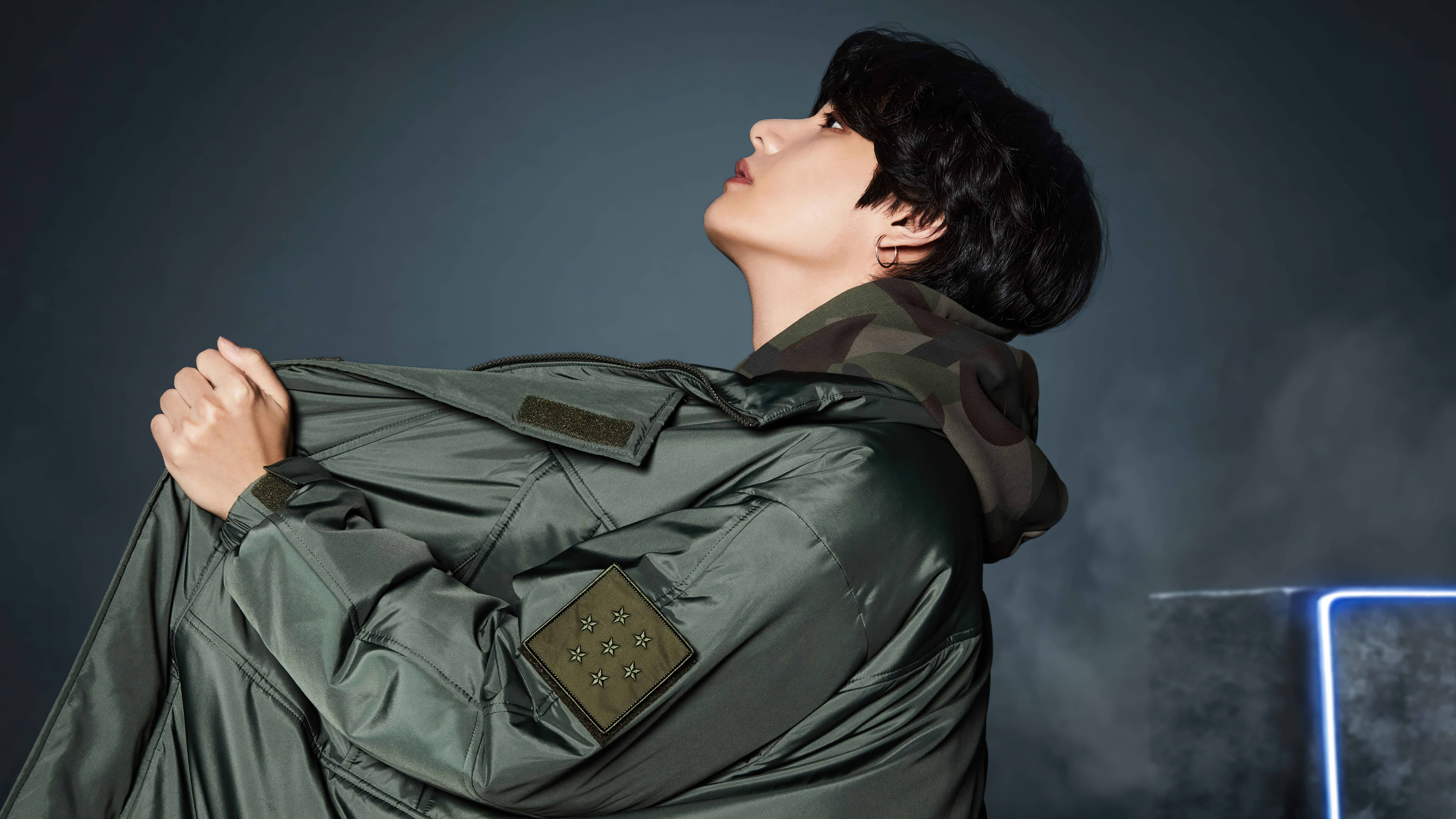 BTS 田柾國 軍綠夾克 壁紙 — 一名身穿綠色軍裝外套的年輕男子，在煙霧瀰漫的黑暗背景中向上看，有一條發光的藍色霓虹線。