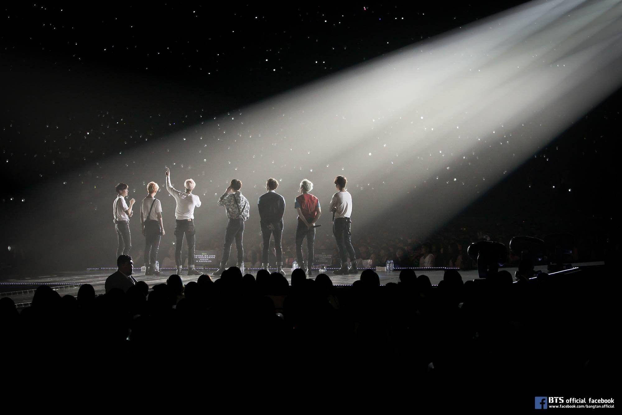 BTS 黑色舞台 壁紙 — 七位表演者站在漆黑的舞台上，沐浴在單一明亮的聚光燈下，背景有星星，觀眾以剪影形式觀看。