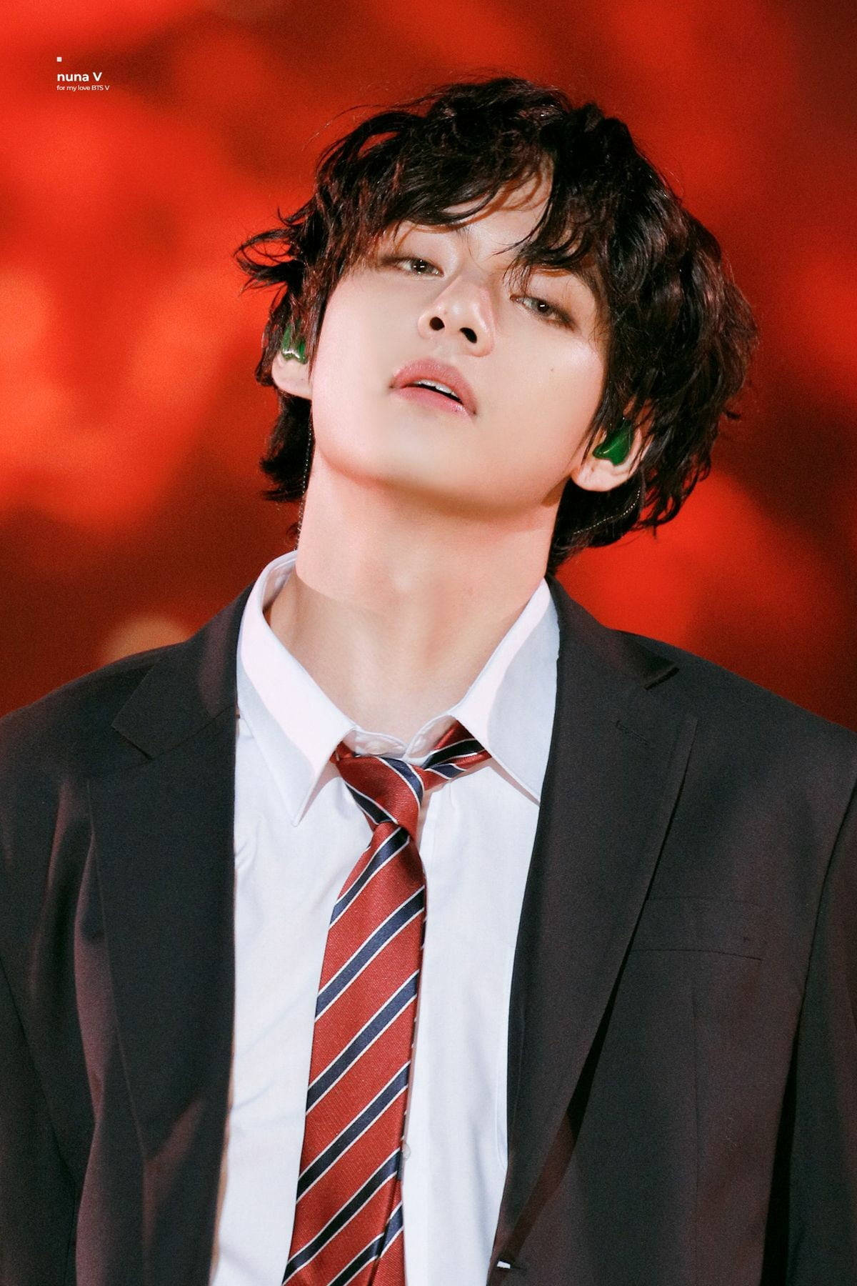 BTS 田柾國銳利眼神 壁紙 — 一名身穿黑色西裝和紅色條紋領帶的年輕男子，在充滿活力的紅色模糊背景前向上凝視。