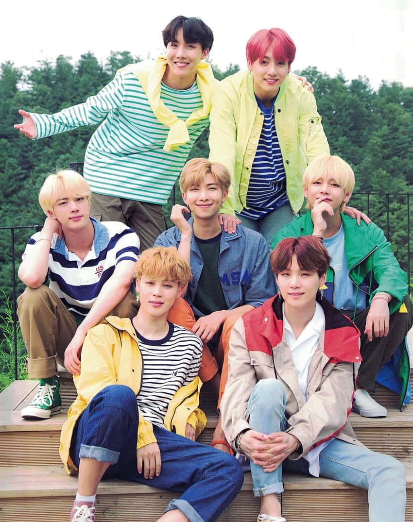 BTS - BTS - BTS - BTS - BTS - B 壁紙 — 七名身穿彩色外套和條紋襯衫的年輕男子在木質台階上愉快地擺姿勢，背景是鬱鬱蔥蔥的綠色森林。