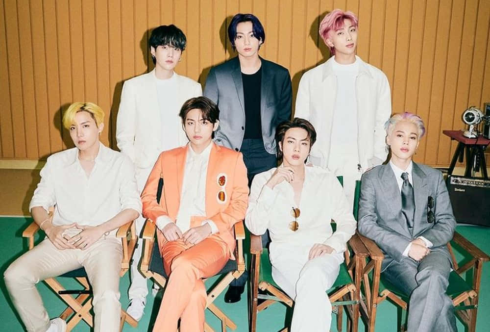 BTS《Butter》照片 壁紙 — 七位穿著時尚、頭髮色彩繽紛的男士自信地坐在導演椅上，背景是溫暖的木鑲板牆。