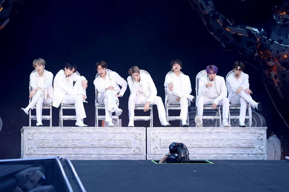BTS《Dionysus》表演後造型 壁紙 — 七位身穿白色西裝的男表演者，在戲劇性的舞台燈光下，隨意地坐在裝飾華麗的椅子上。