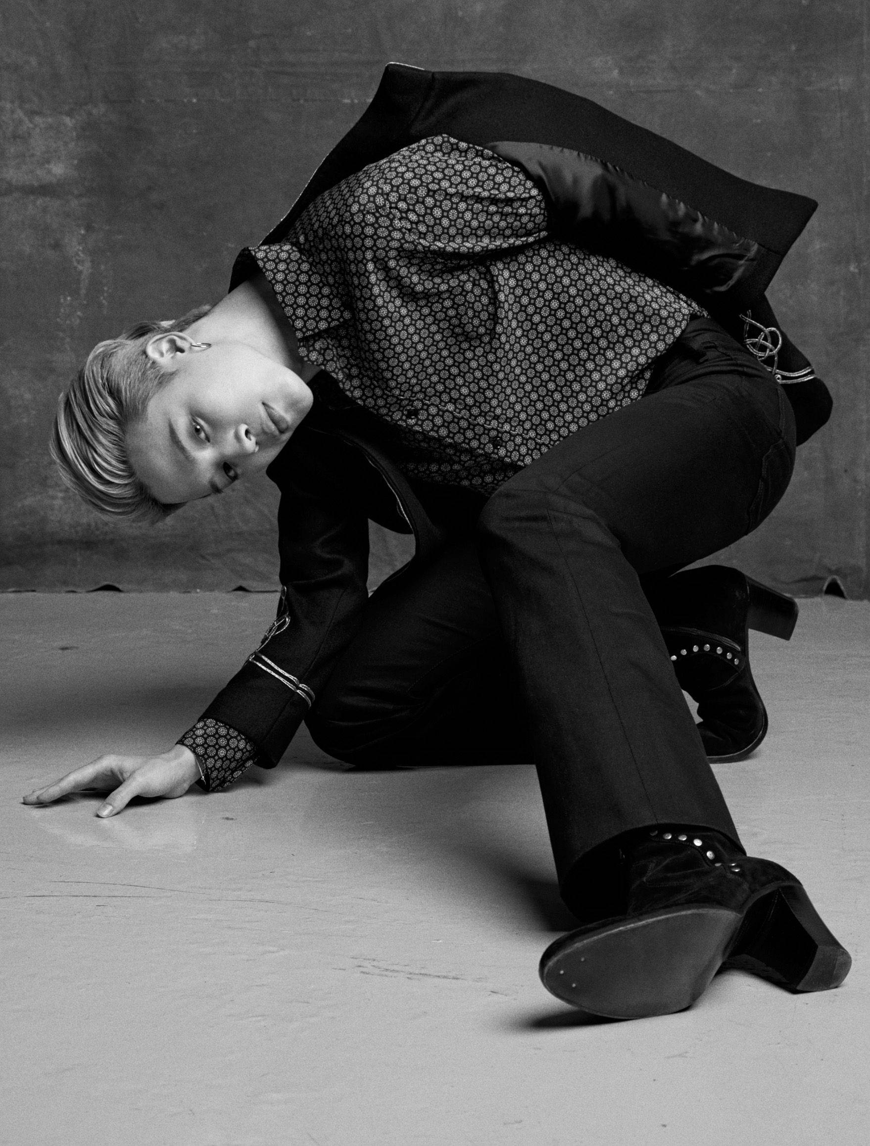 BTS 朴智旻 Esquire雜誌 壁紙 — 一個穿著圖案襯衫和黑色褲子的人，在單色調的紋理灰色牆壁前，在混凝土地板上動態擺姿勢。