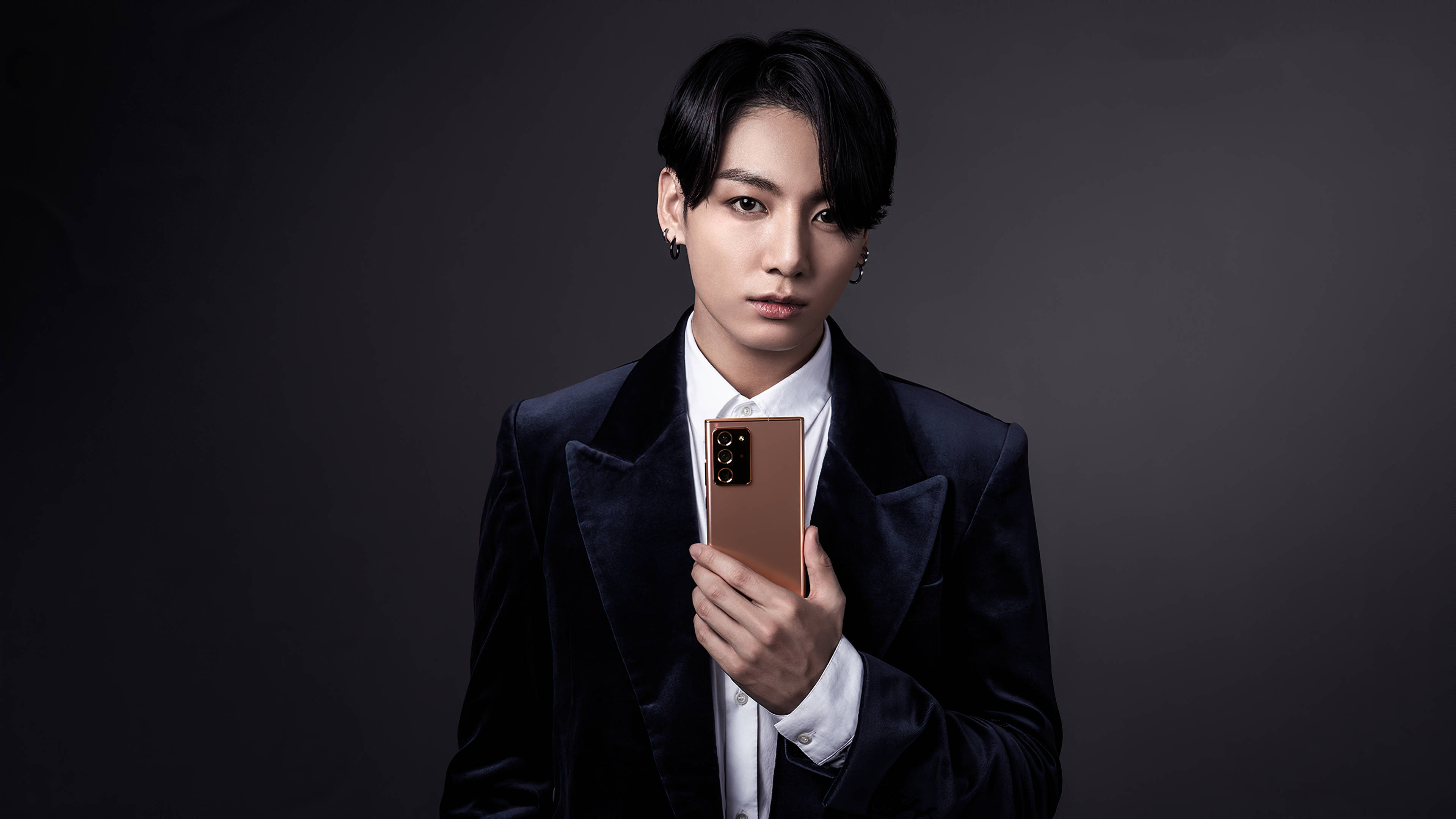 BTS 田柾國手機模特兒美學照 壁紙 — 一名身穿深色天鵝絨西裝的男子，在柔和的灰色背景前手持玫瑰金手機，眼神直接而強烈。