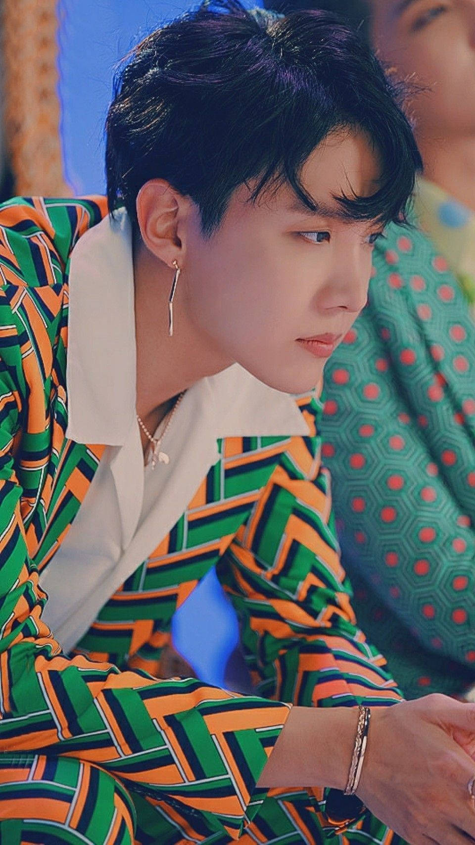 BTS Idol J-hope 壁紙 — 一位深色頭髮的年輕男子穿著大膽的綠橙條紋襯衫和銀飾，若有所思地看向一旁。
