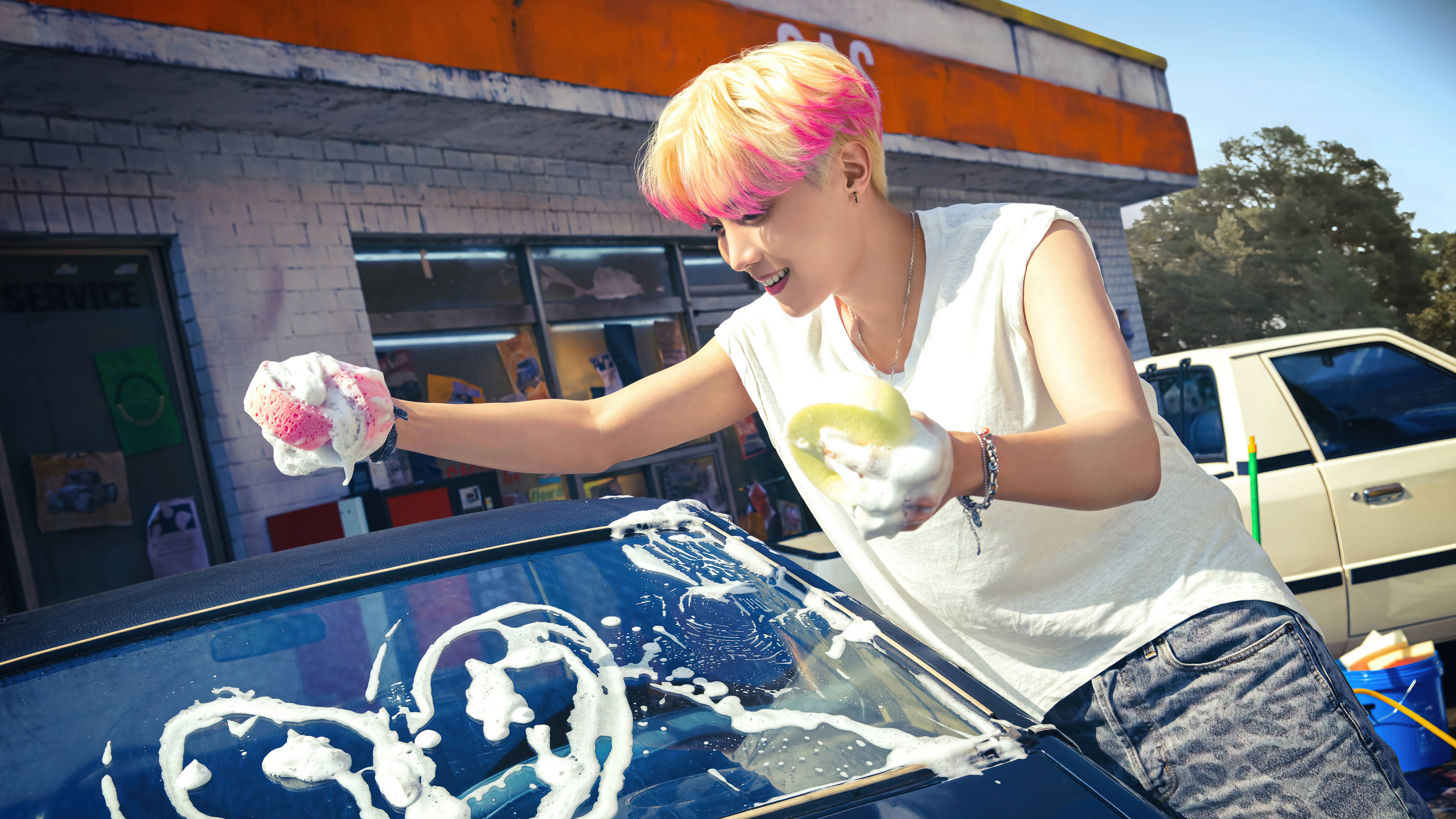 BTS J-hope 洗車男孩 壁紙 — 一個擁有鮮豔粉紅色和金色頭髮的人，在明亮的日光下愉快地用肥皂泡沫洗車。