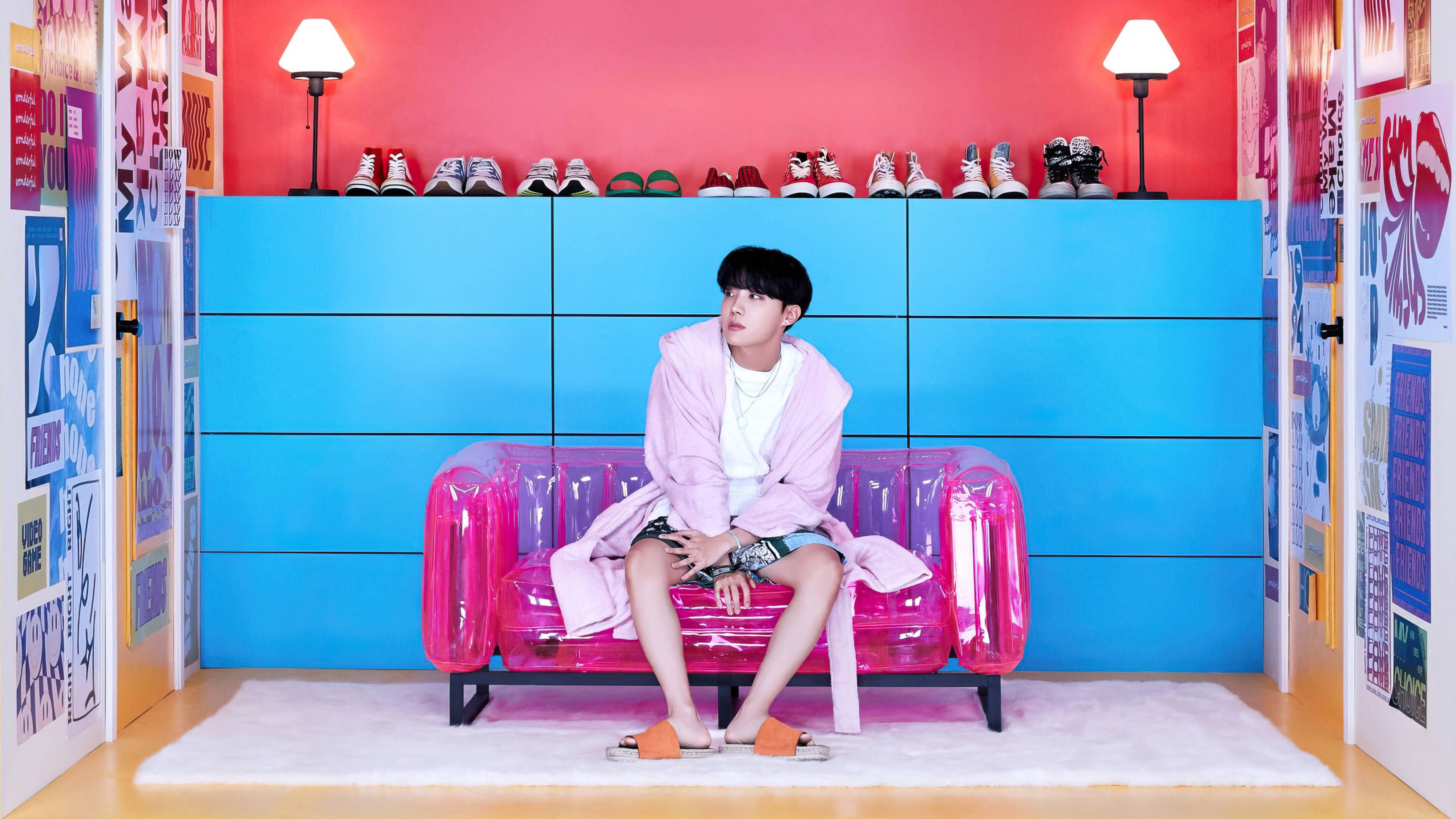 BTS J-hope BE專輯 壁紙 — 一個年輕男子坐在粉紅色充氣沙發上，房間充滿活力，藍色牆壁和彩色海報。