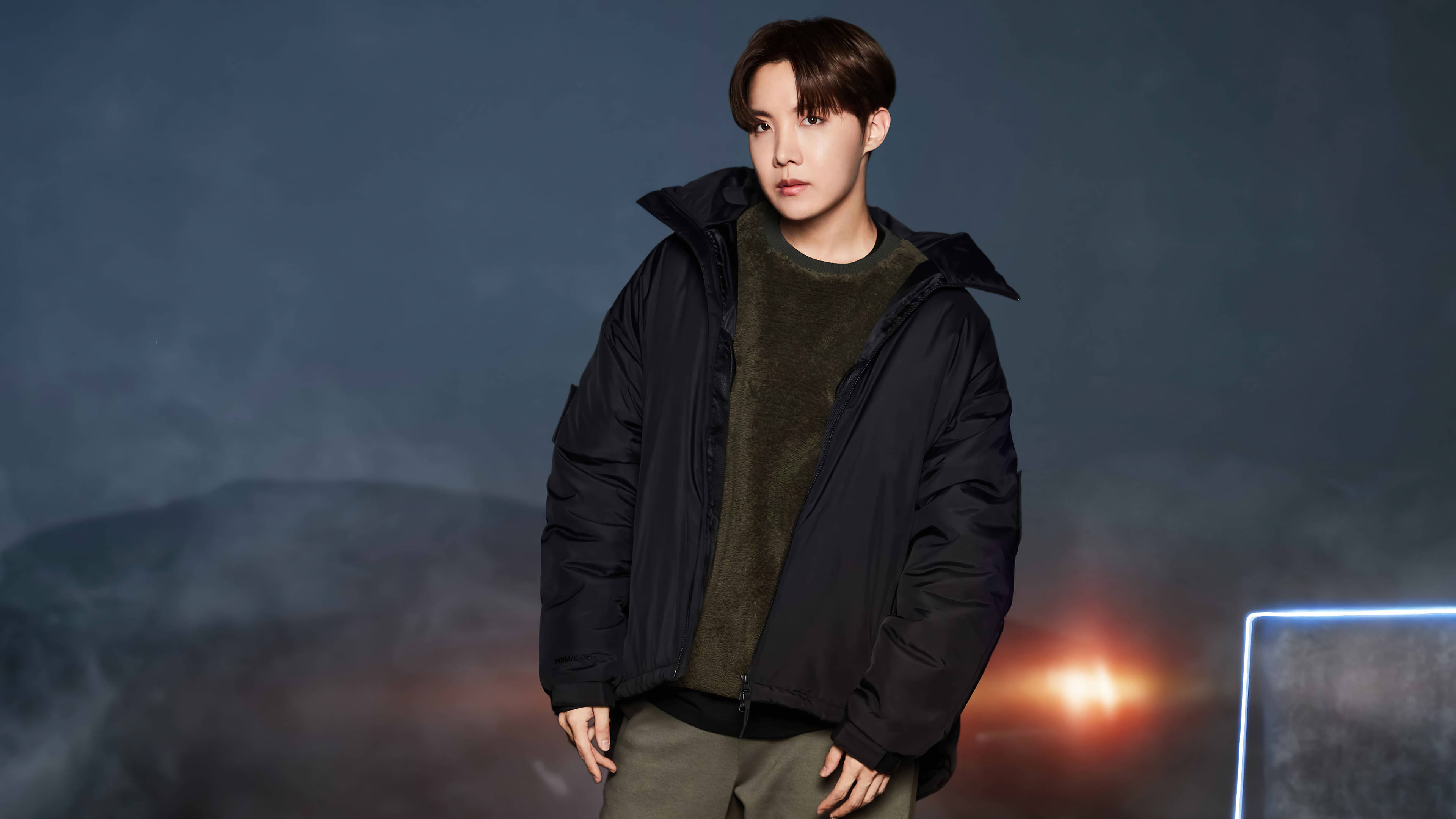 BTS J-hope FILA拍攝 壁紙 — 一位身穿黑色羽絨外套的年輕男子自信地站在一個帶有發光橙色燈光和未來感藍色霓虹燈的陰暗背景下。