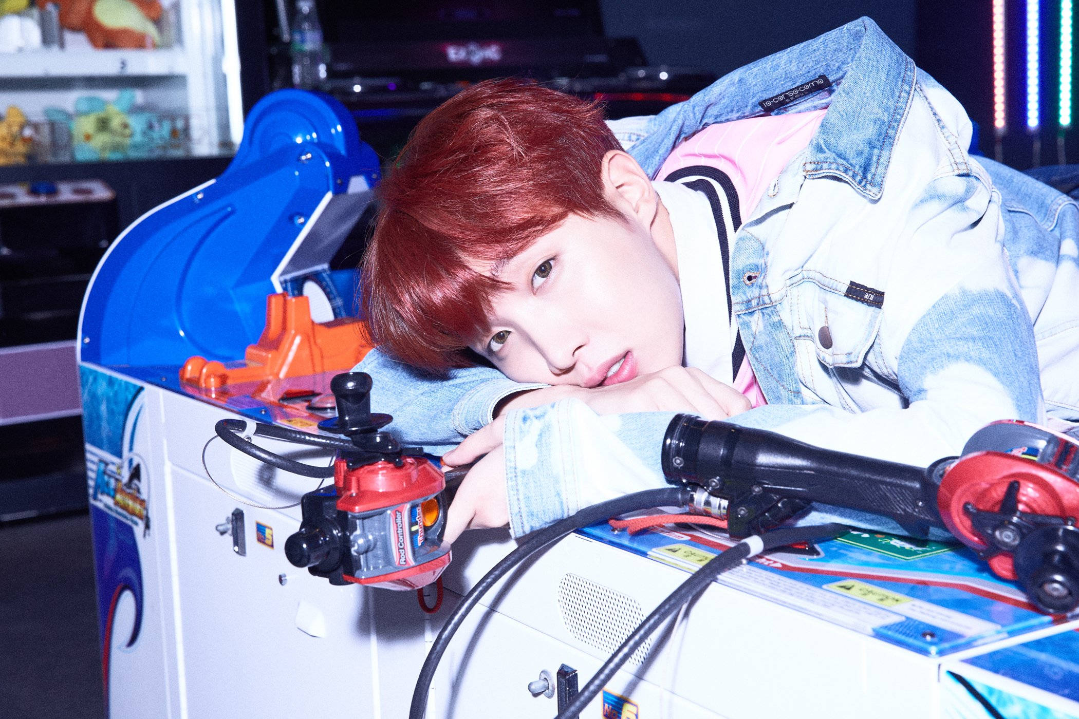 BTS J-hope In Arcade 壁紙 — 一位紅髮年輕男子在昏暗的遊戲室裡俯身於色彩繽紛的街機上。
