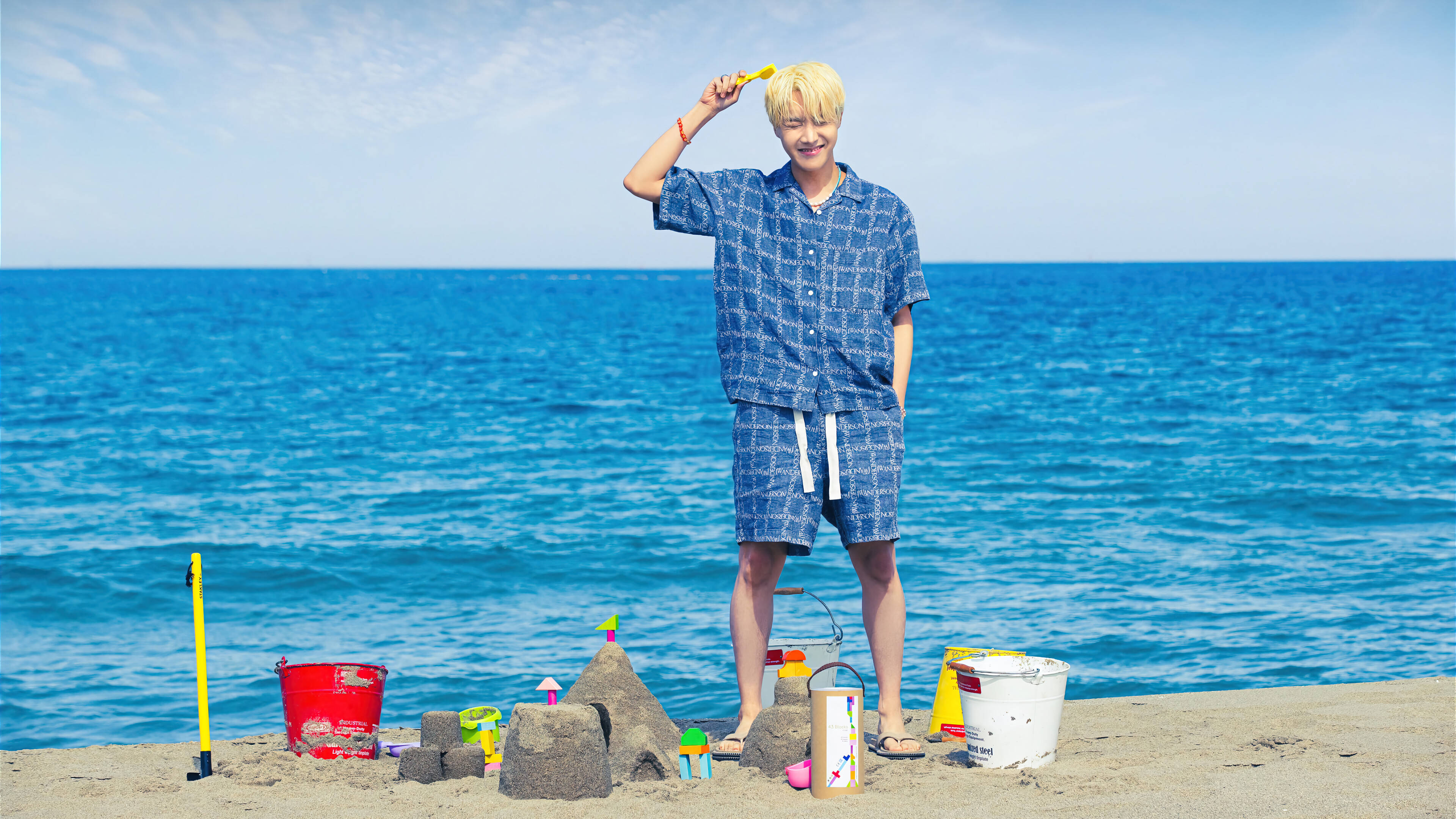 BTS J-hope 海灘 壁紙 — 一位金髮年輕男子站在陽光明媚的海灘上，拿著黃色鏟子，旁邊有沙堡和色彩繽紛的沙灘玩具。