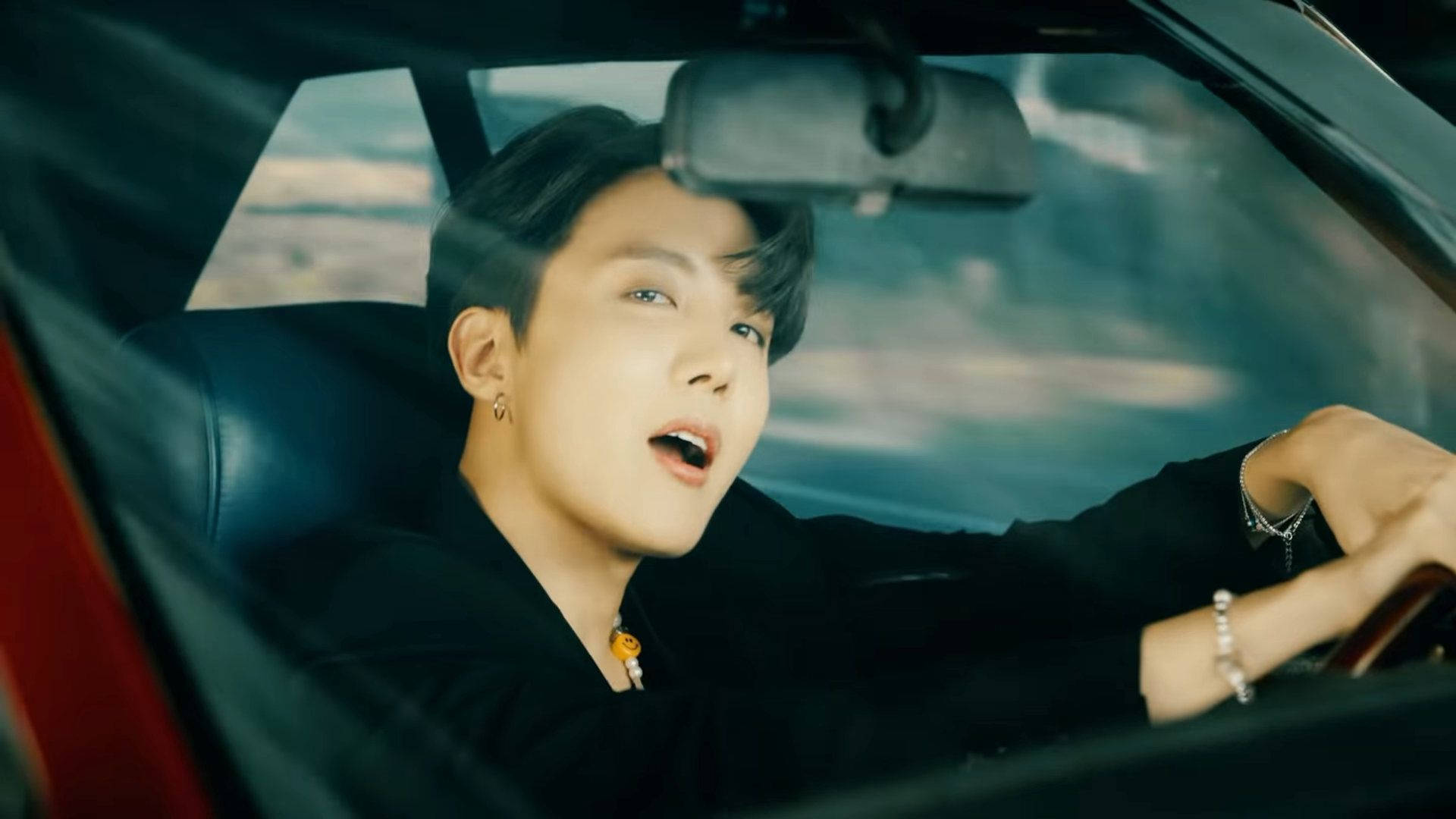 BTS J-hope 駕車 壁紙 — 一位黑髮戴耳環的年輕男子張開嘴開車，身穿黑色衣服和銀色手鐲。