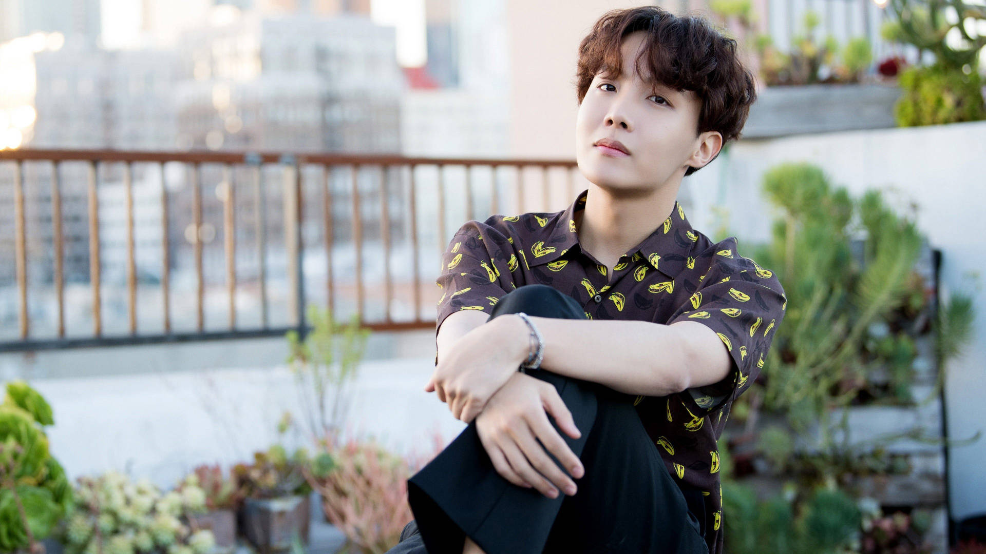 BTS J-Hope戶外休閒照 壁紙 — 一個捲髮年輕人在黃金時刻坐在屋頂花園，周圍有盆栽植物和城市建築，若有所思。