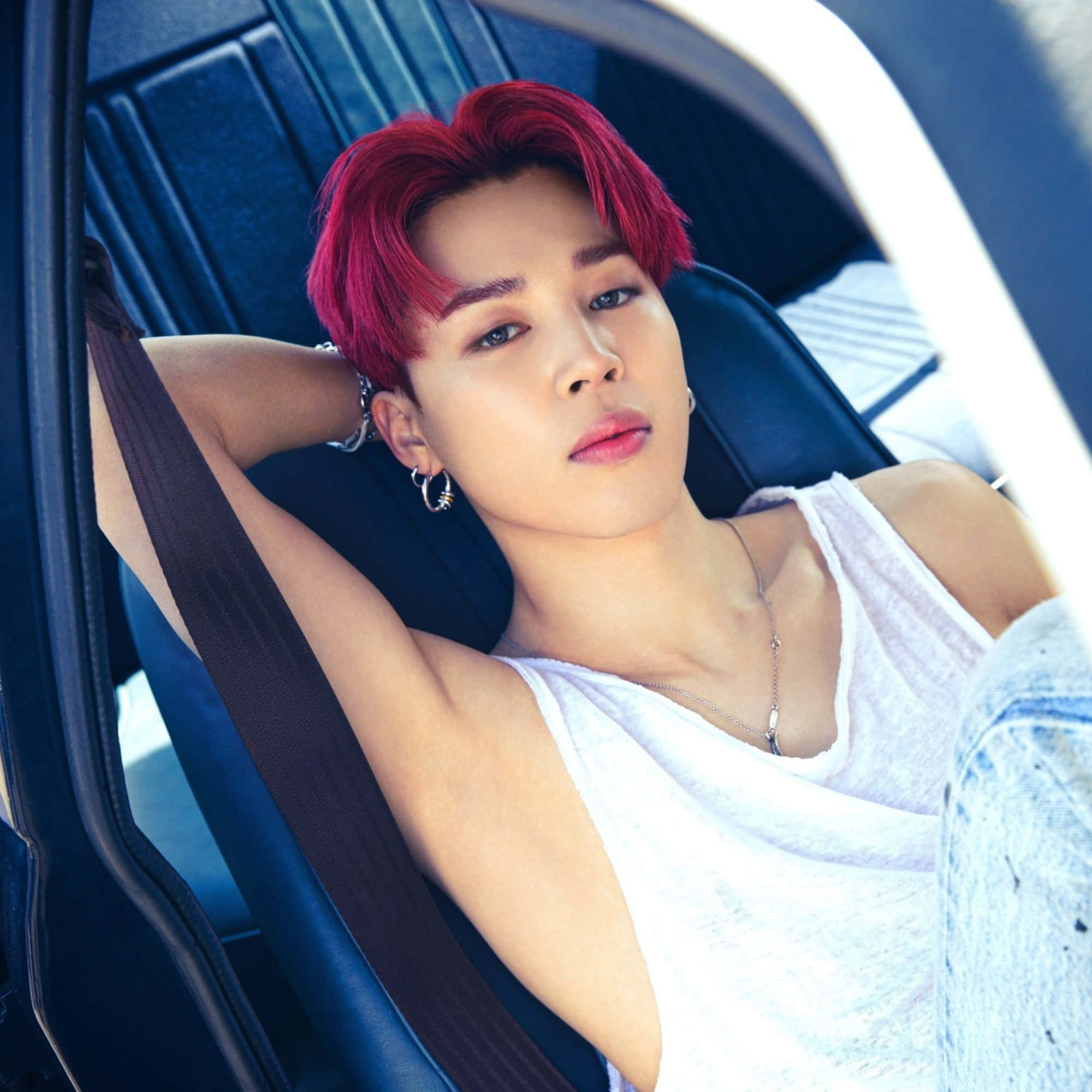 BTS 的 Jimin 看起來帥氣逼人 壁紙 — 一位擁有鮮豔紅髮的年輕男子自信地靠在汽車座椅上，身穿白色背心和牛仔褲。