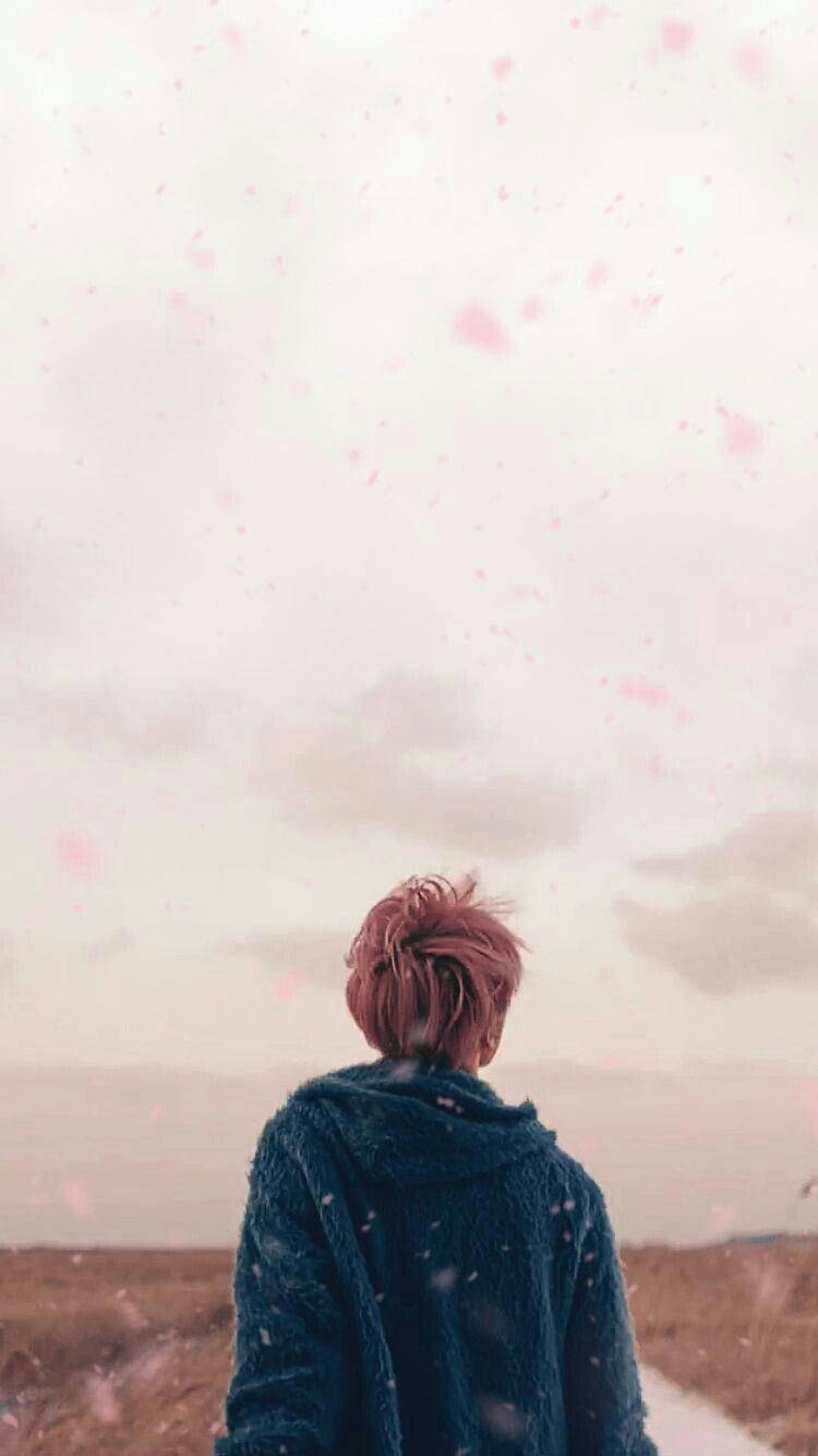 粉紅色頭髮、穿著藍色連帽衫的人站在粉紅色花瓣天空下的田野中