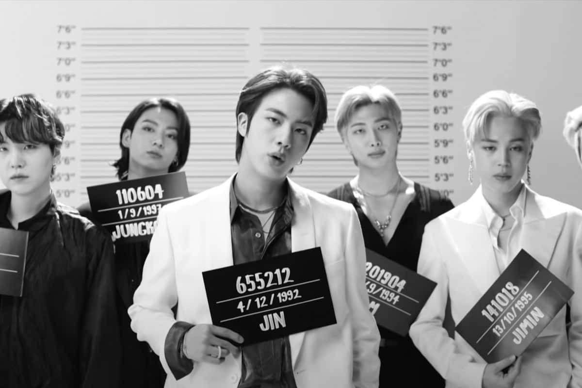 BTS Jin 黑白監獄風寫真 壁紙 — 黑白風格的大頭照肖像，五名身穿西裝的男子在身高圖表背景前手持身份證。