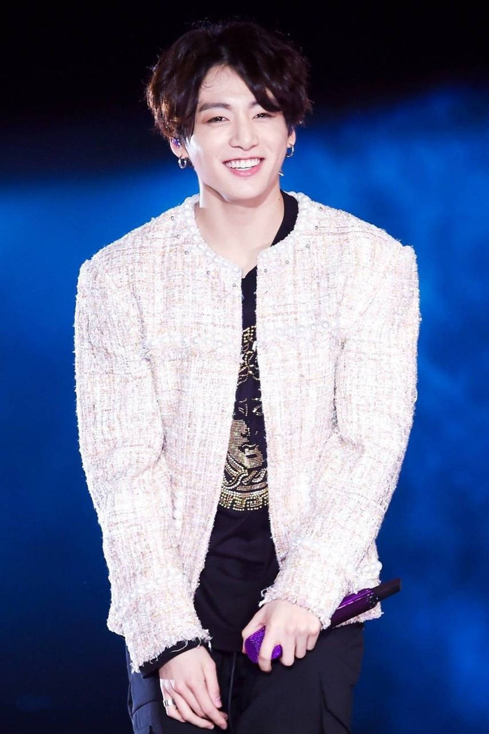 BTS JK 可愛舞台表演 壁紙 — 一位微笑的表演者穿著質感白色外套和黑色襯衫，在藍色舞台燈光下拿著紫色麥克風。
