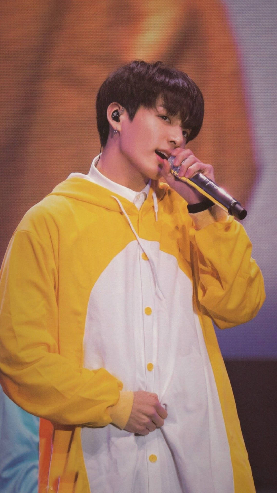 BTS JK第四次粉絲見面會演出服裝 壁紙 — 一位身穿黃白連帽衫的年輕男子在溫暖的舞台燈光下對著麥克風唱歌。